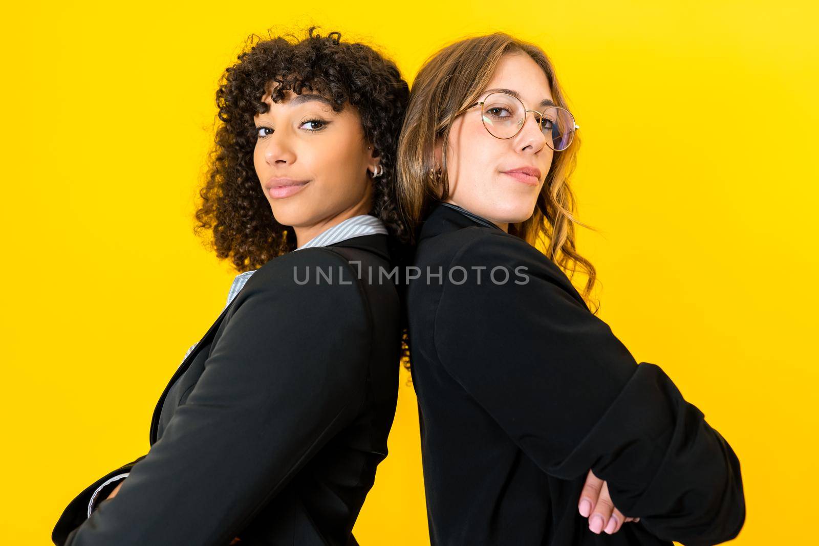 Confident multiracial business women back to back with crossed arms looking at camera isolated on yellow background for copy space. Female success on work and better life concept by robbyfontanesi
