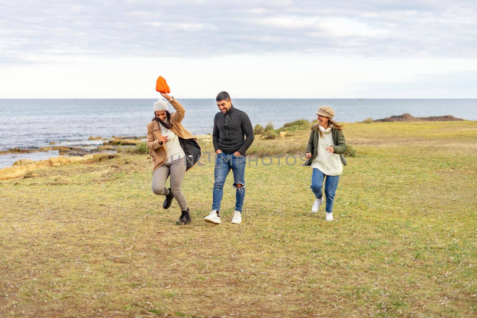 Three mixed-race carefree friends running outdoor joking each other - Millennial people having fun on a meadow near sea smiling in winter vacation enjoying nature in ocean resort wearing clothing by robbyfontanesi