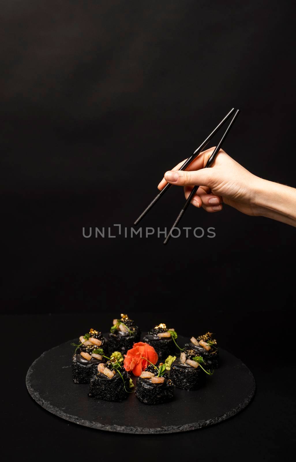 Hand with chopsticks wants to take custom sushi roll with black rice, crab meat, avocado, smoked salmon mousse, oar caviar, masago, shrimp cocktail, edible gold leaf, ginger, wasabi on black table