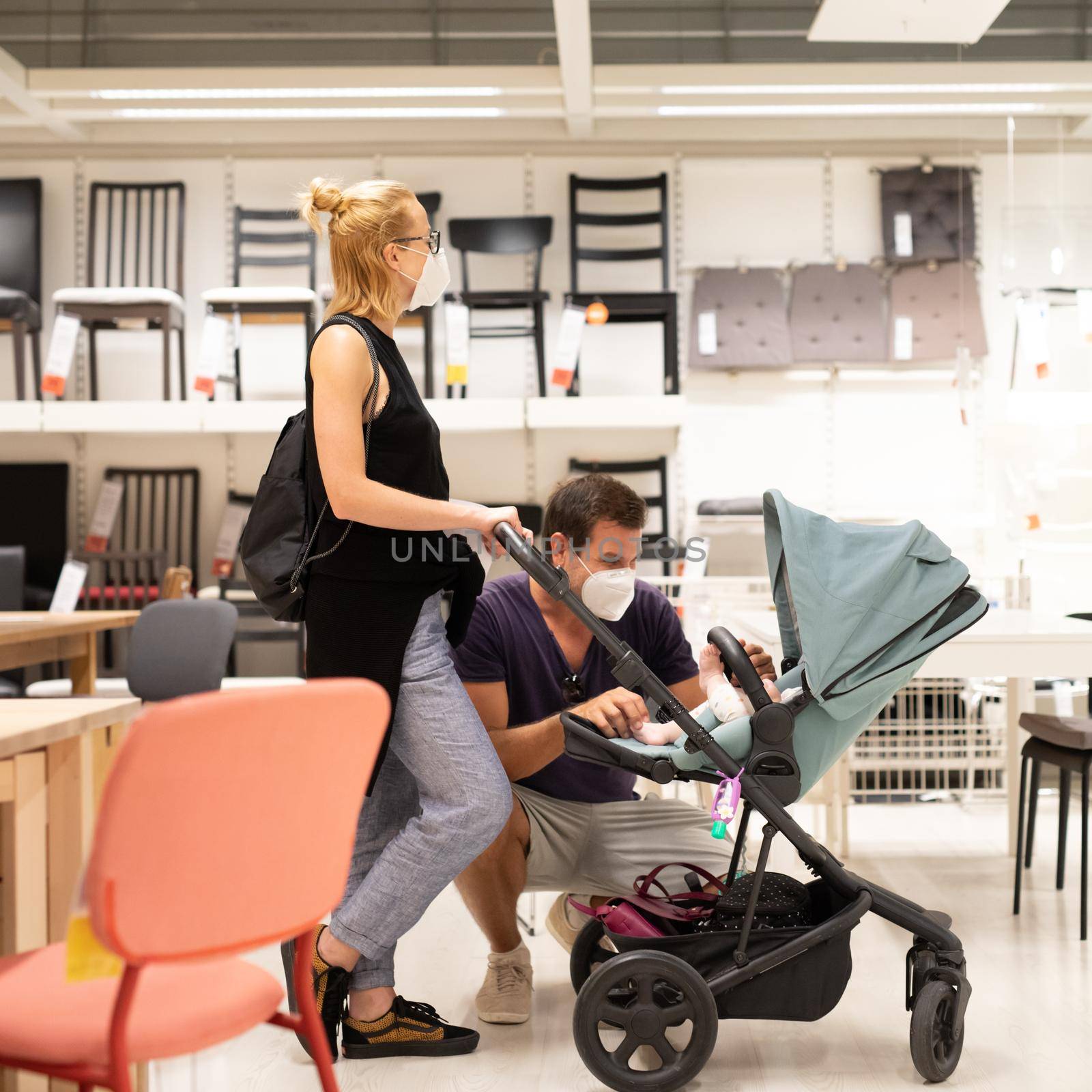 Young family with newborn in stroller shopping at retail furniture and home accessories store wearing protective medical face mask to prevent spreading of corona virus when shops reopen.