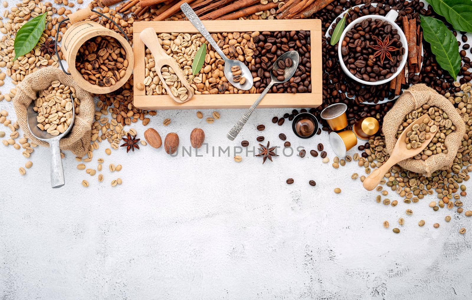 Green and brown decaf unroasted and dark roasted coffee beans in wooden box with scoops setup on white concrete background.