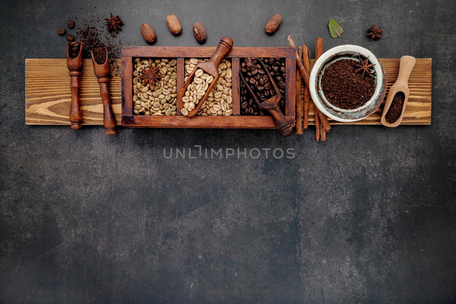 Green and brown unroasted and dark roasted coffee beans in wooden box with scoops setup on dark stone background.