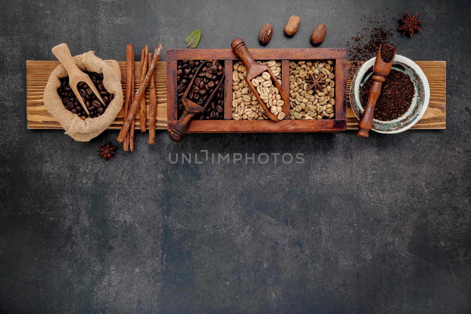 Green and brown unroasted and dark roasted coffee beans in wooden box with scoops setup on dark stone background.