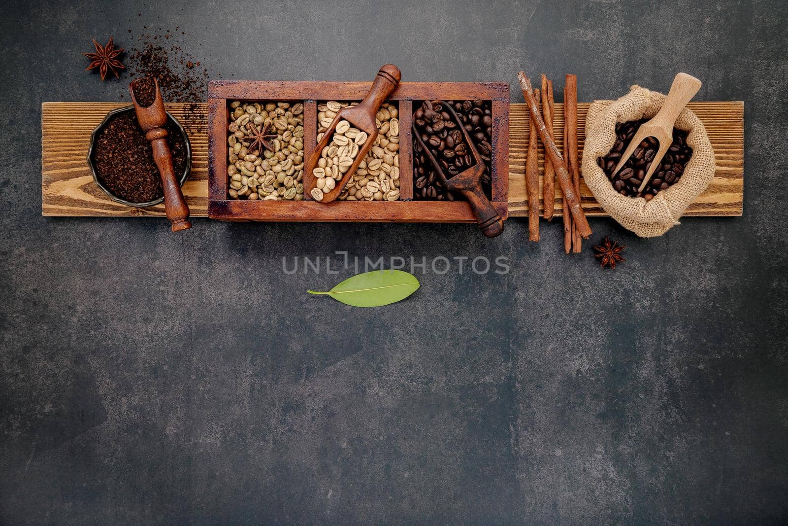 Green and brown unroasted and dark roasted coffee beans in wooden box with scoops setup on dark stone background.