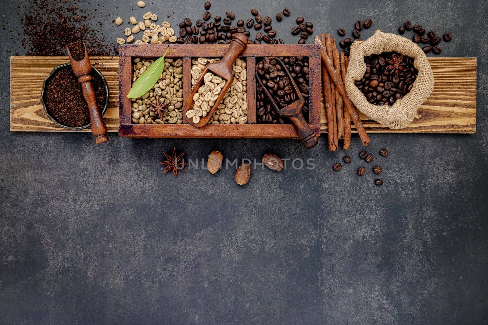 Green and brown unroasted and dark roasted coffee beans in wooden box with scoops setup on dark stone background.