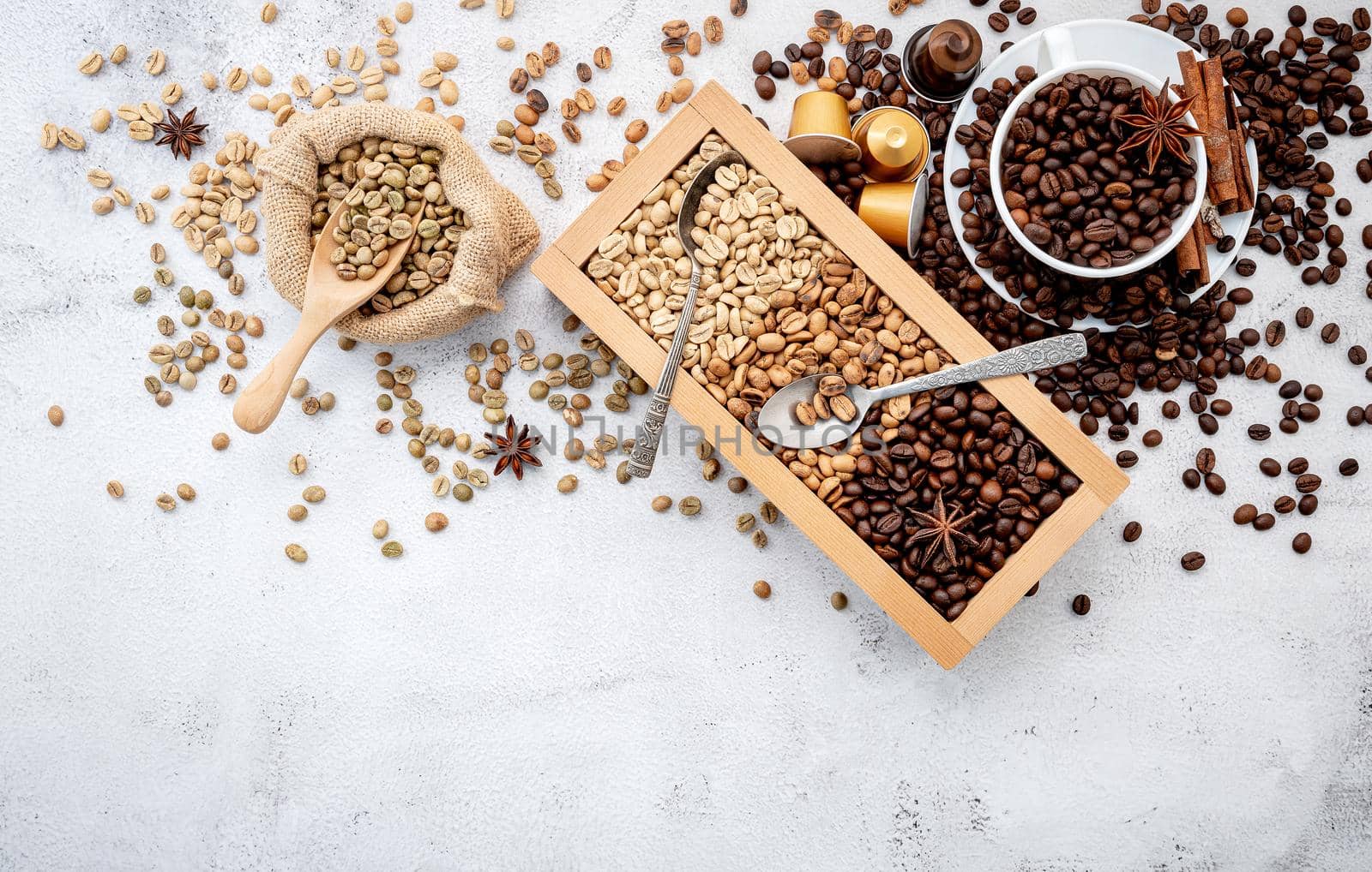 Green and brown decaf unroasted and dark roasted coffee beans in wooden box with scoops setup on white concrete background.