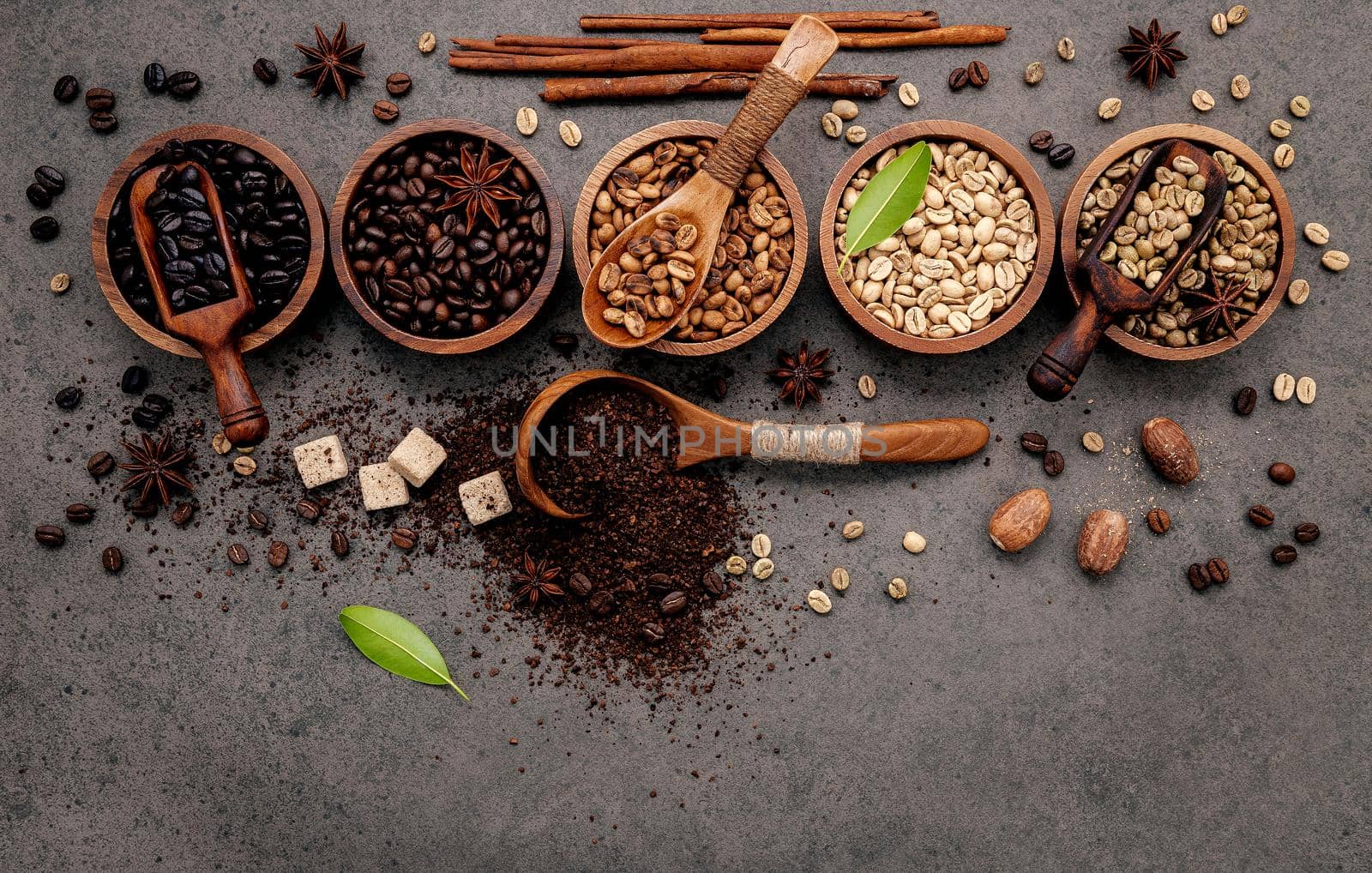 Green and brown unroasted and dark roasted coffee beans in wooden bowl with spoons set up on dark concrete background.