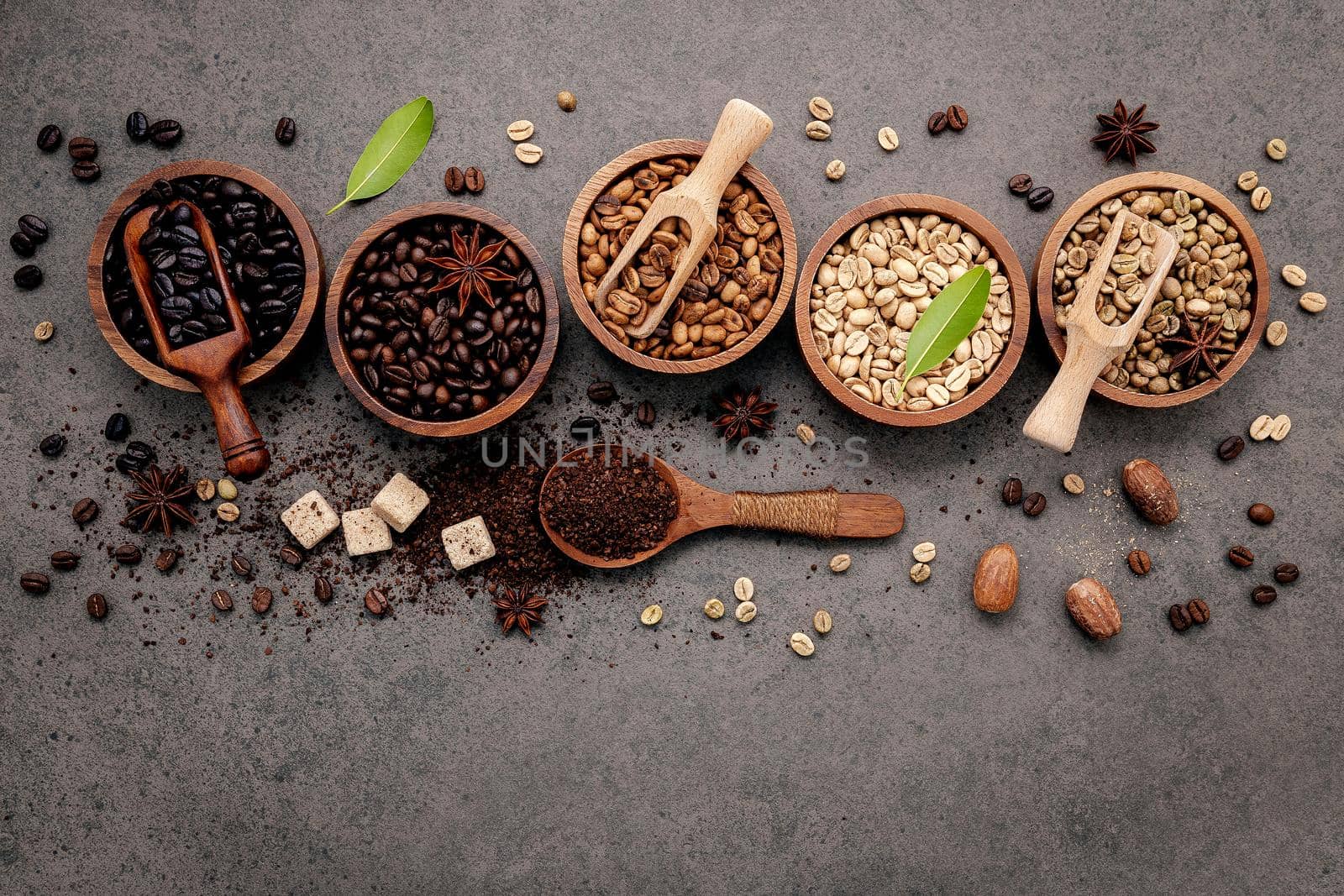 Green and brown unroasted and dark roasted coffee beans in wooden bowl with spoons set up on dark concrete background.