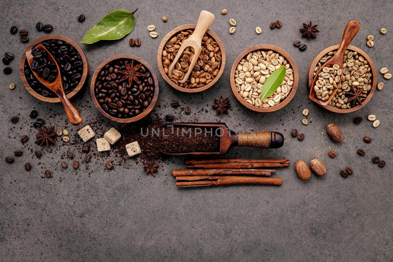 Green and brown unroasted and dark roasted coffee beans in wooden bowl with spoons set up on dark concrete background.