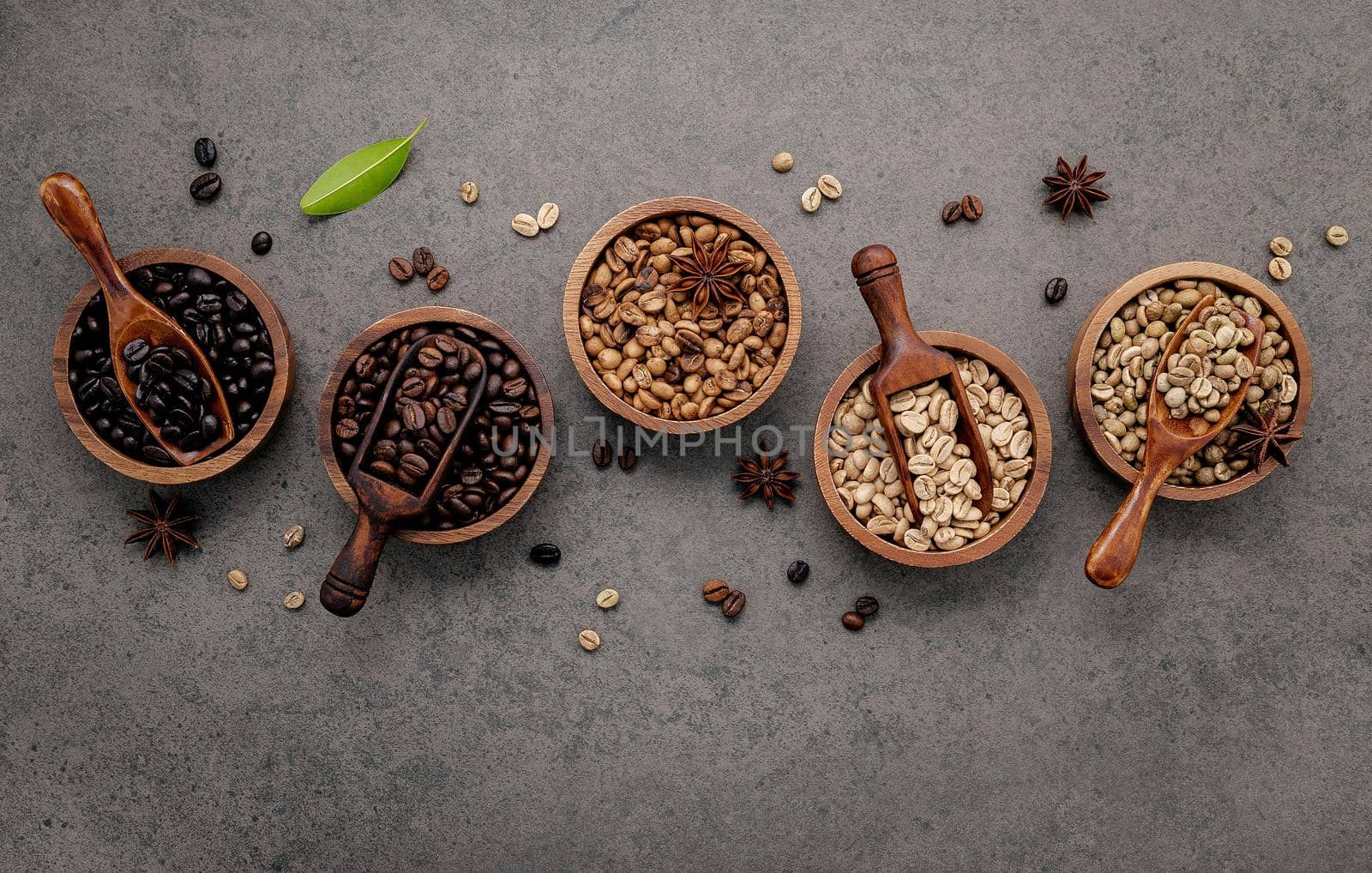 Green and brown unroasted and dark roasted coffee beans in wooden bowl with spoons set up on dark concrete background.