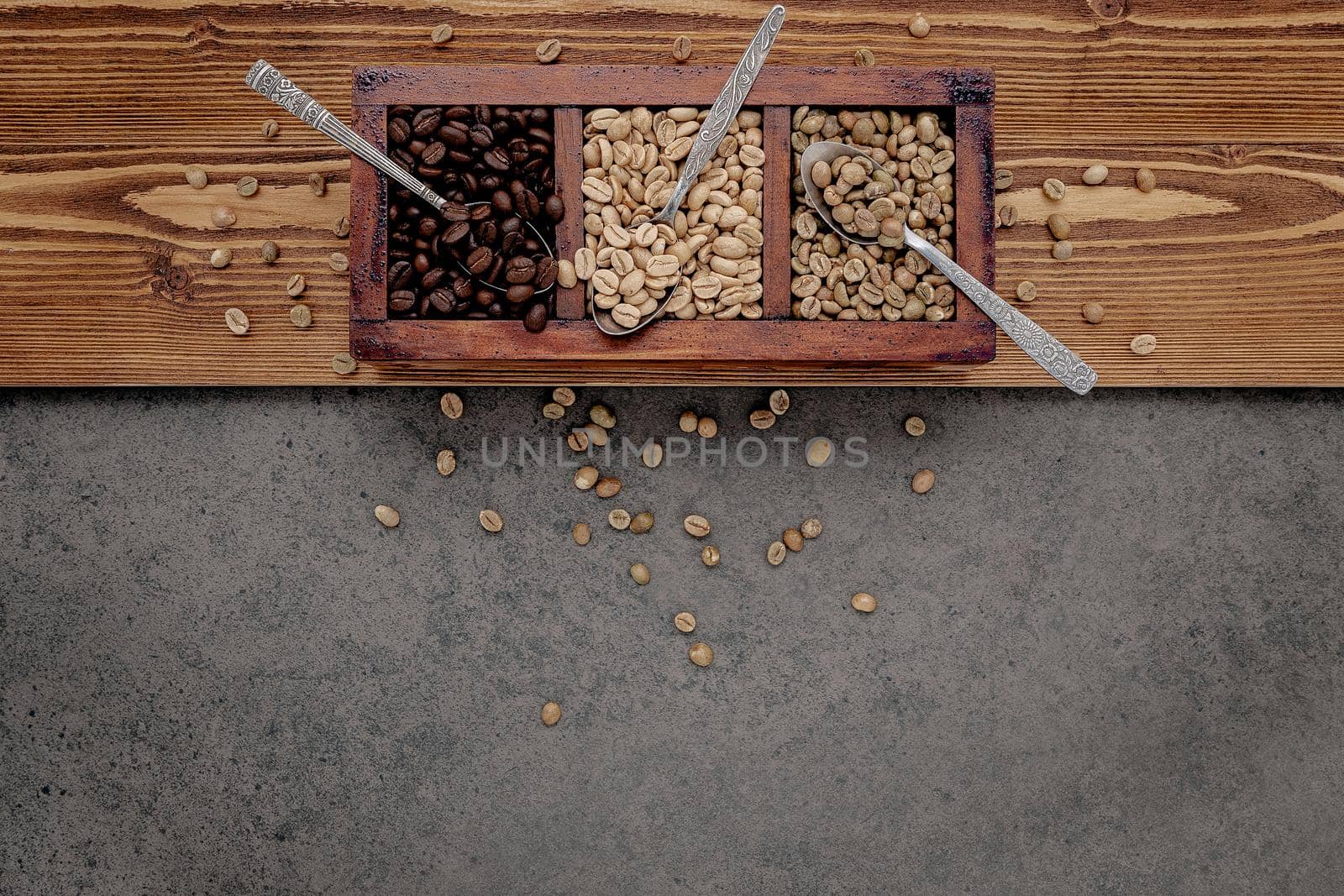 Green and brown unroasted and dark roasted coffee beans in wooden box with spoons setup on dark concrete background.