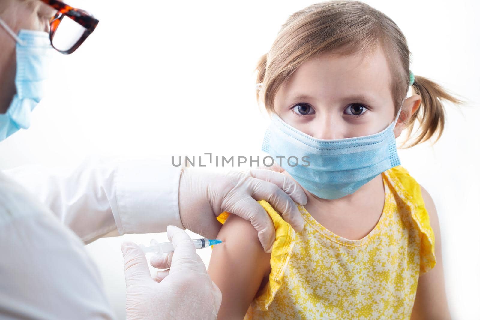 Vaccination concept in the era of coronavirus. Woman Doctor vaccinating cute little girl wearing yellow dress and facial protective mask on white background.