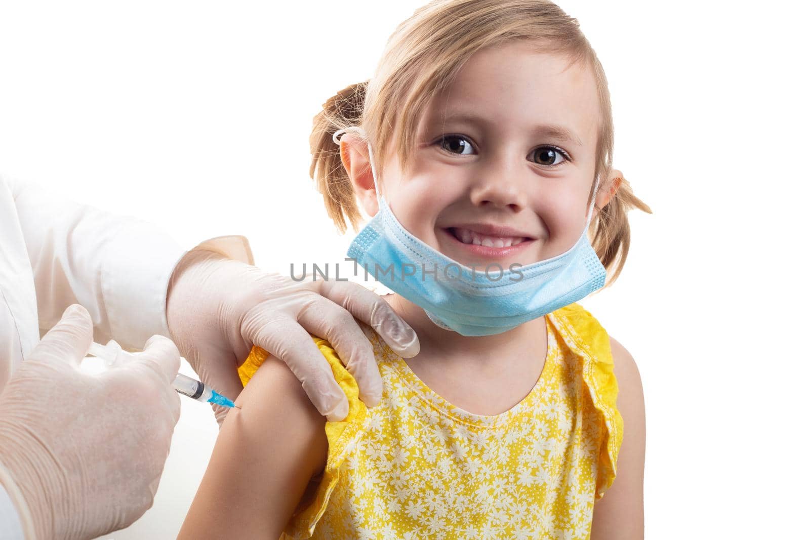 Vaccination concept in the era of coronavirus. Doctor vaccinating cute smiling little girl wearing yellow dress and facial protective mask on white background.