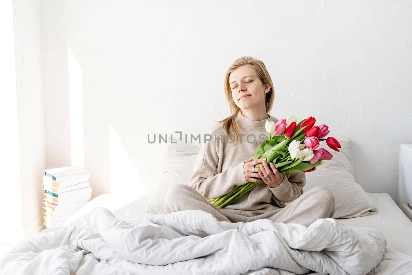 Happy woman sitting on the bed wearing pajamas holding tulip flowers bouquet with eyes closed by Desperada