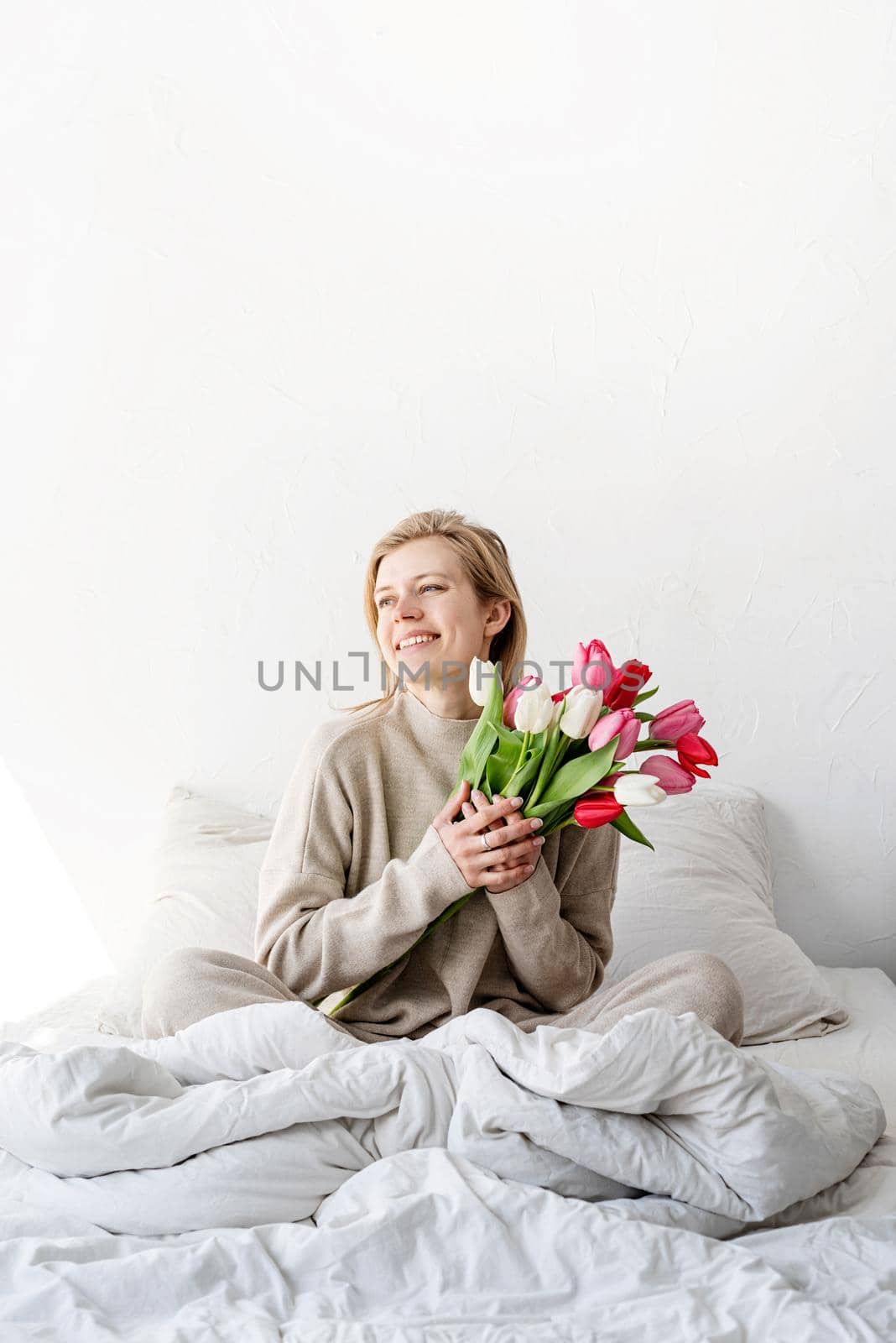 Happy woman sitting on the bed wearing pajamas holding tulip flowers bouquet by Desperada