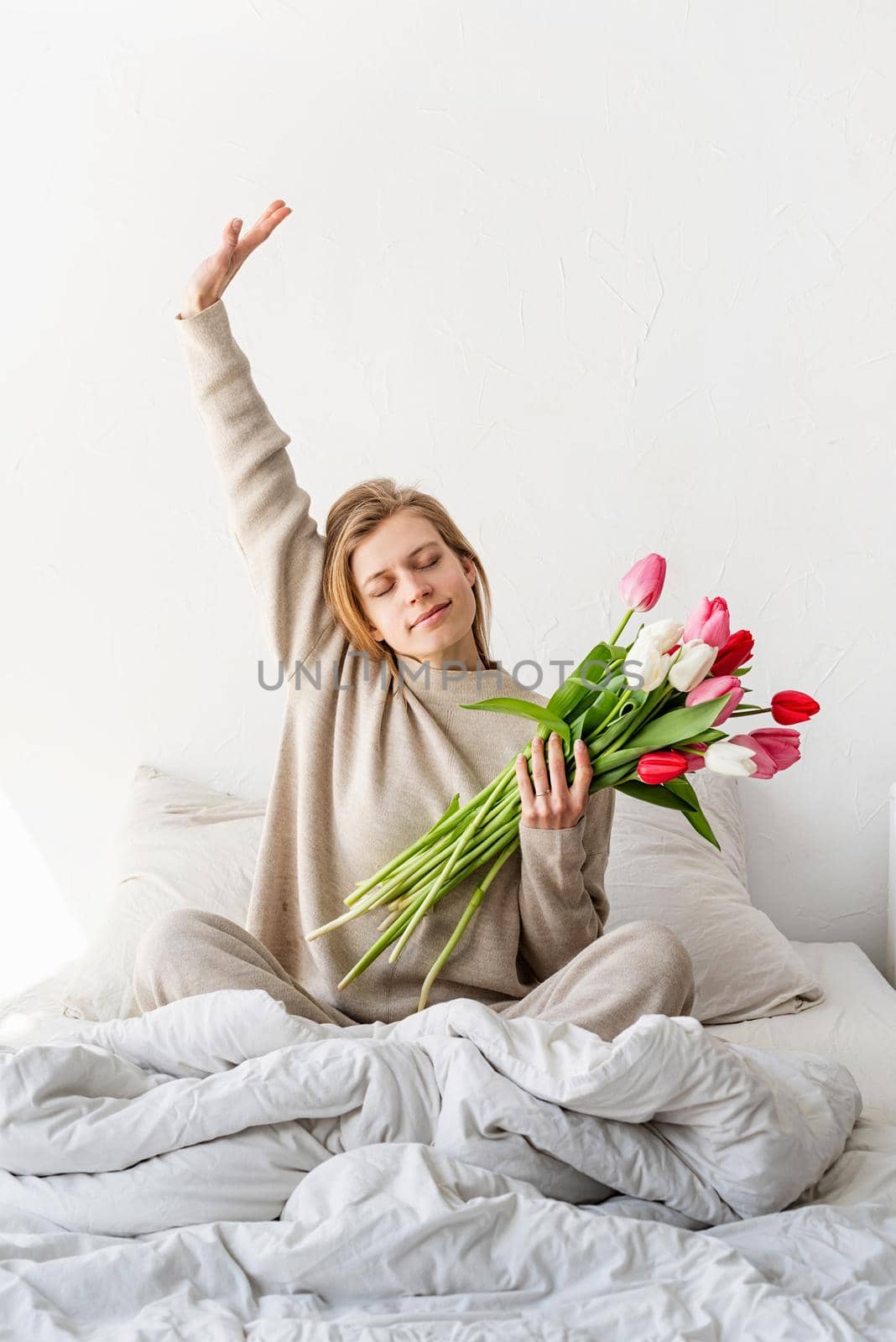 Happy woman sitting on the bed wearing pajamas stretching and holding tulip flowers bouquet by Desperada