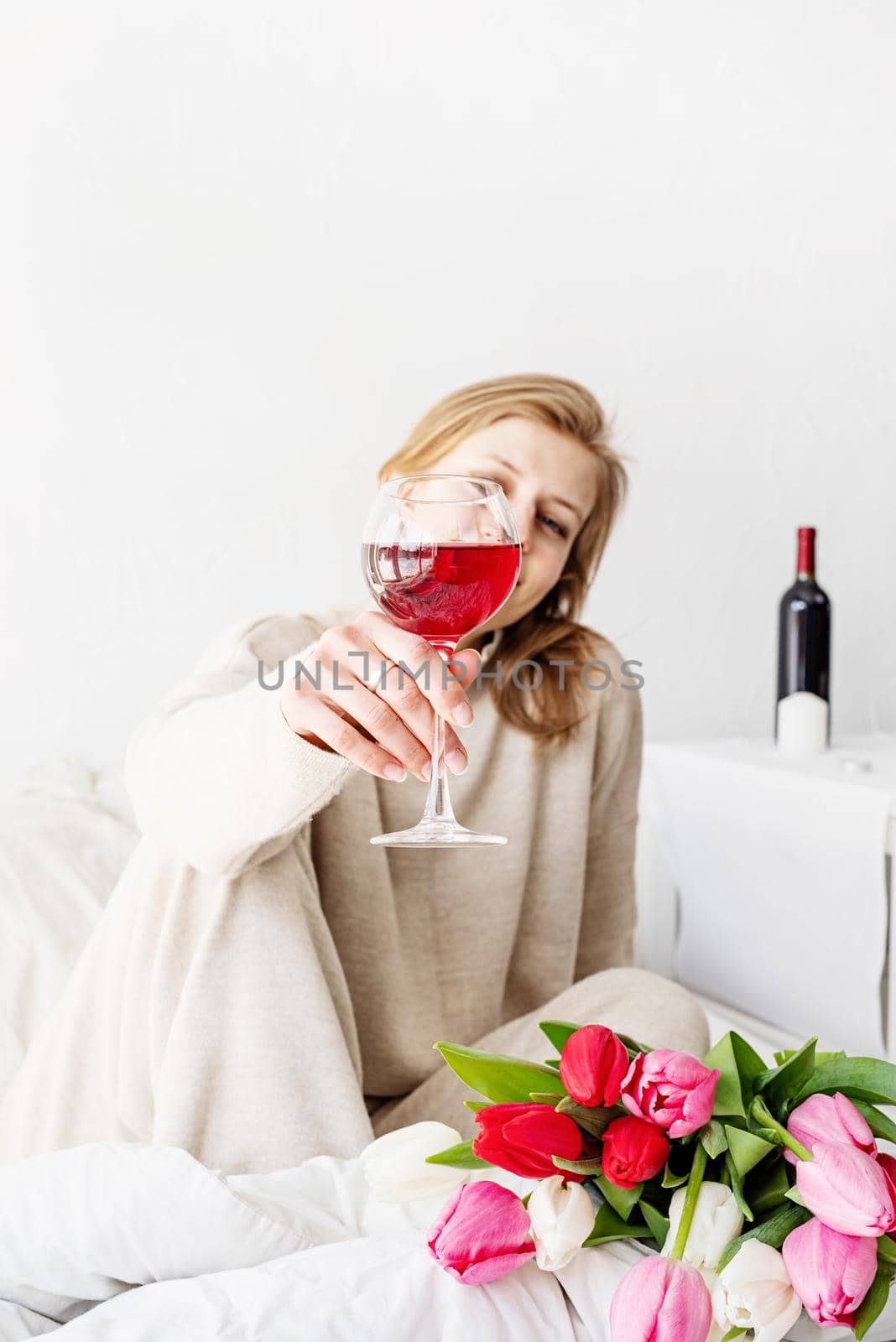 Happy woman sitting on the bed wearing pajamas, with pleasure enjoying flowers and a glass of red wine