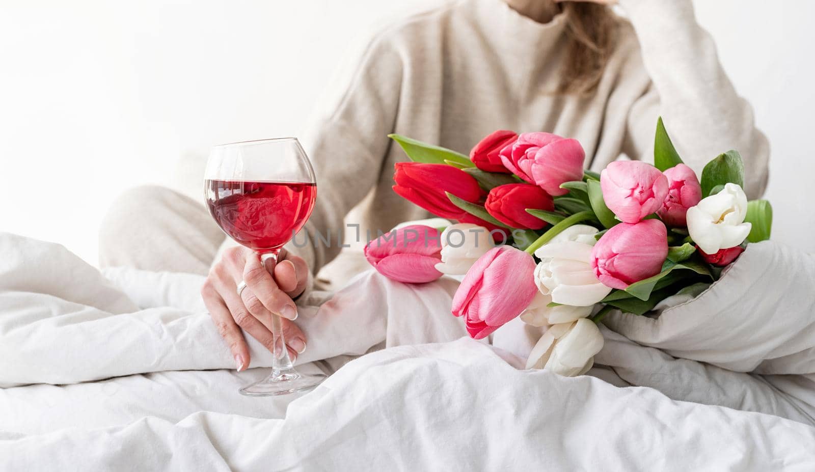 Happy woman sitting on the bed wearing pajamas, with pleasure enjoying flowers and a glass of red wine