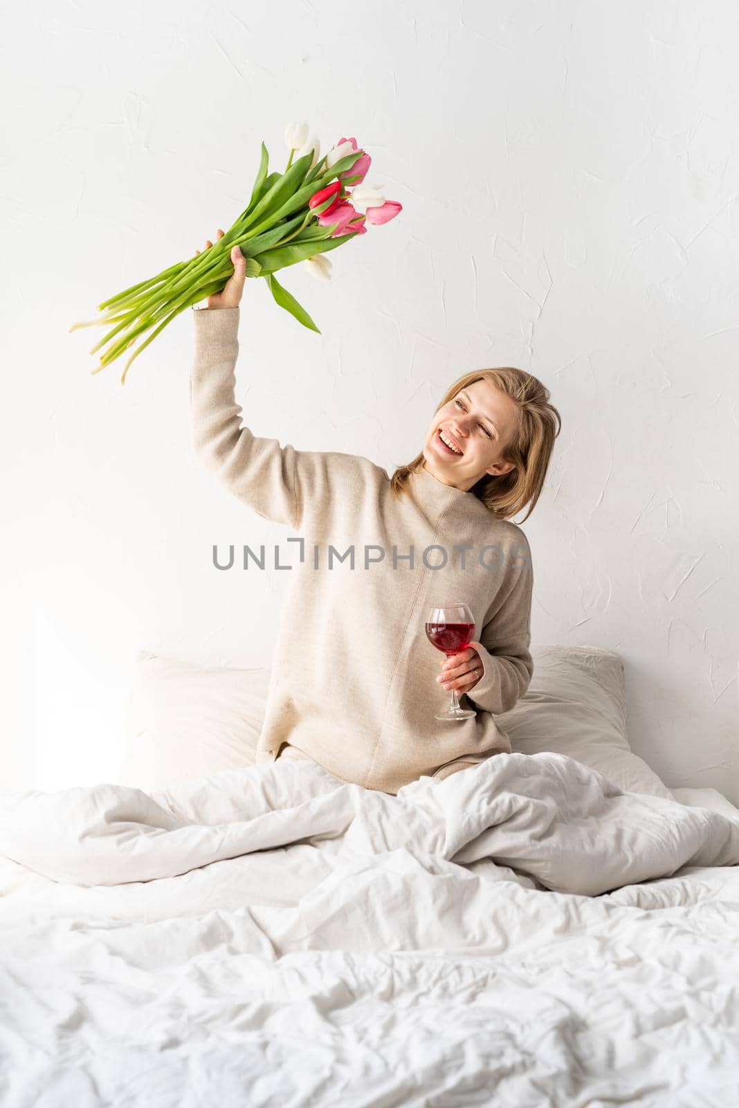 Happy woman sitting on the bed wearing pajamas holding glass of wine and tulip flowers bouquet by Desperada