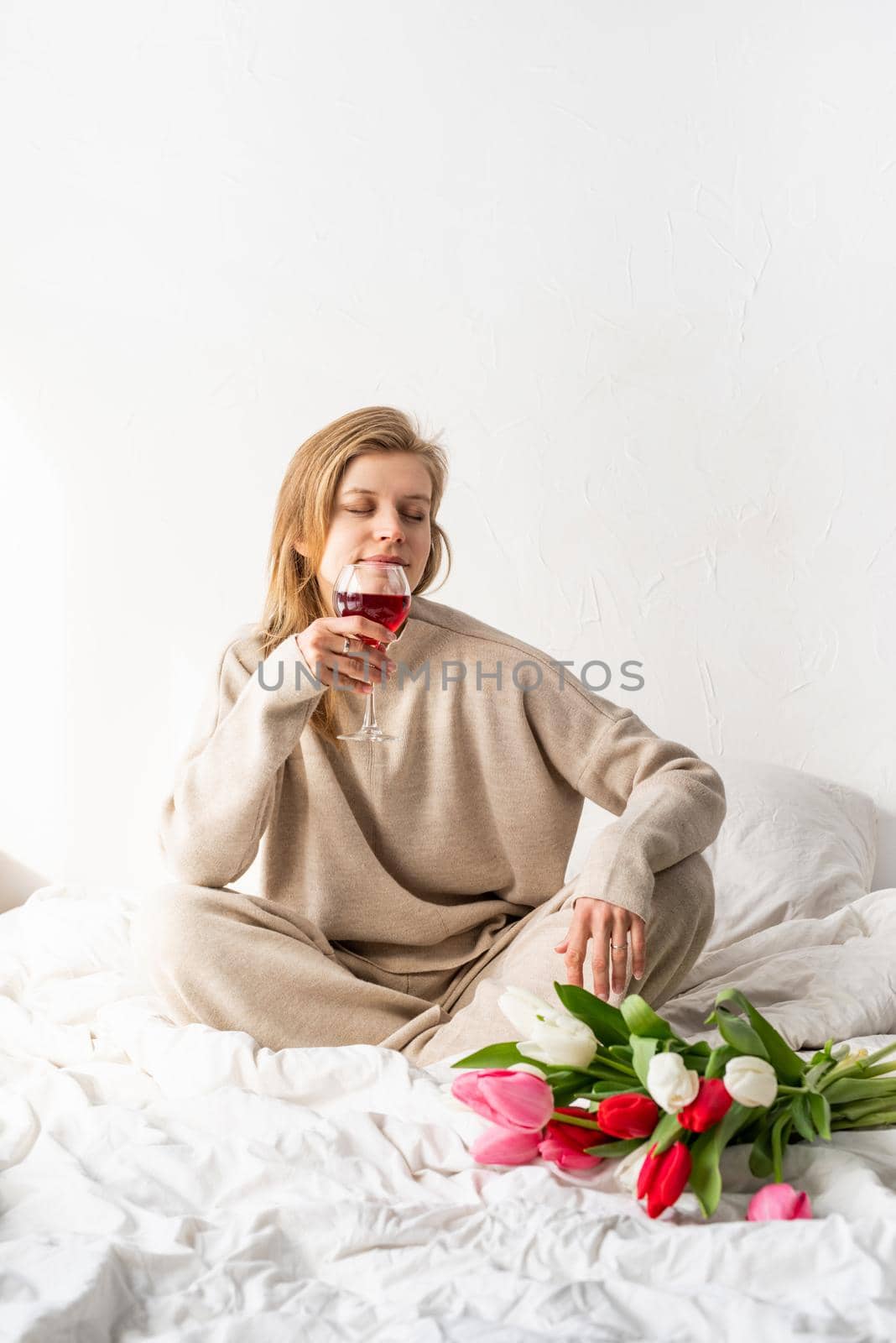 Happy woman sitting on the bed wearing pajamas, with pleasure enjoying flowers and a glass of red wine