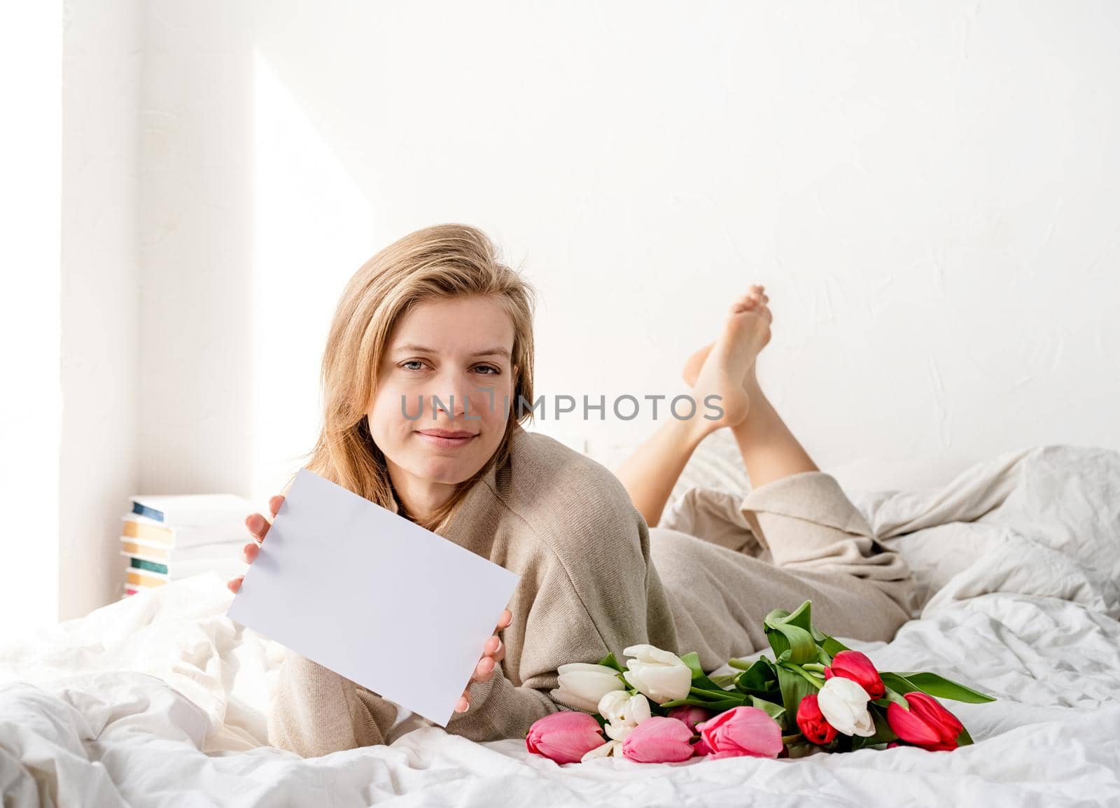 Happy woman lying on the bed wearing pajamas, holding tulip flowers bouquet and blank card for mock up design