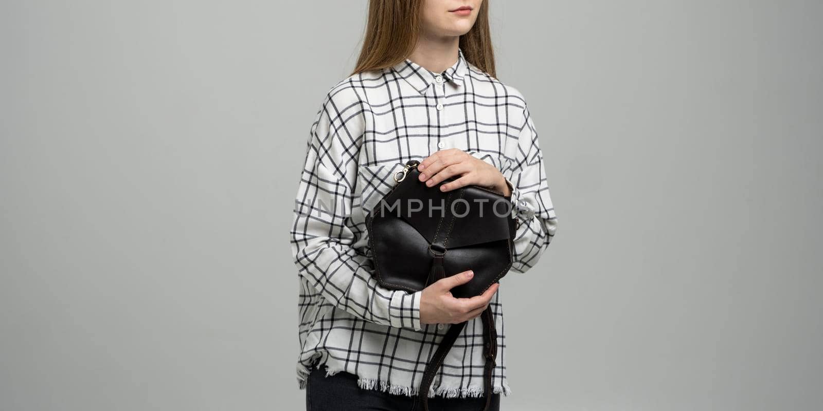 Small black leather bag in a woman's hand on a white background. Shoulder handbag. Woman in a white plaid shirt and black jeans and with a black handbag. Style, retro, fashion, vintage and elegance. by vovsht