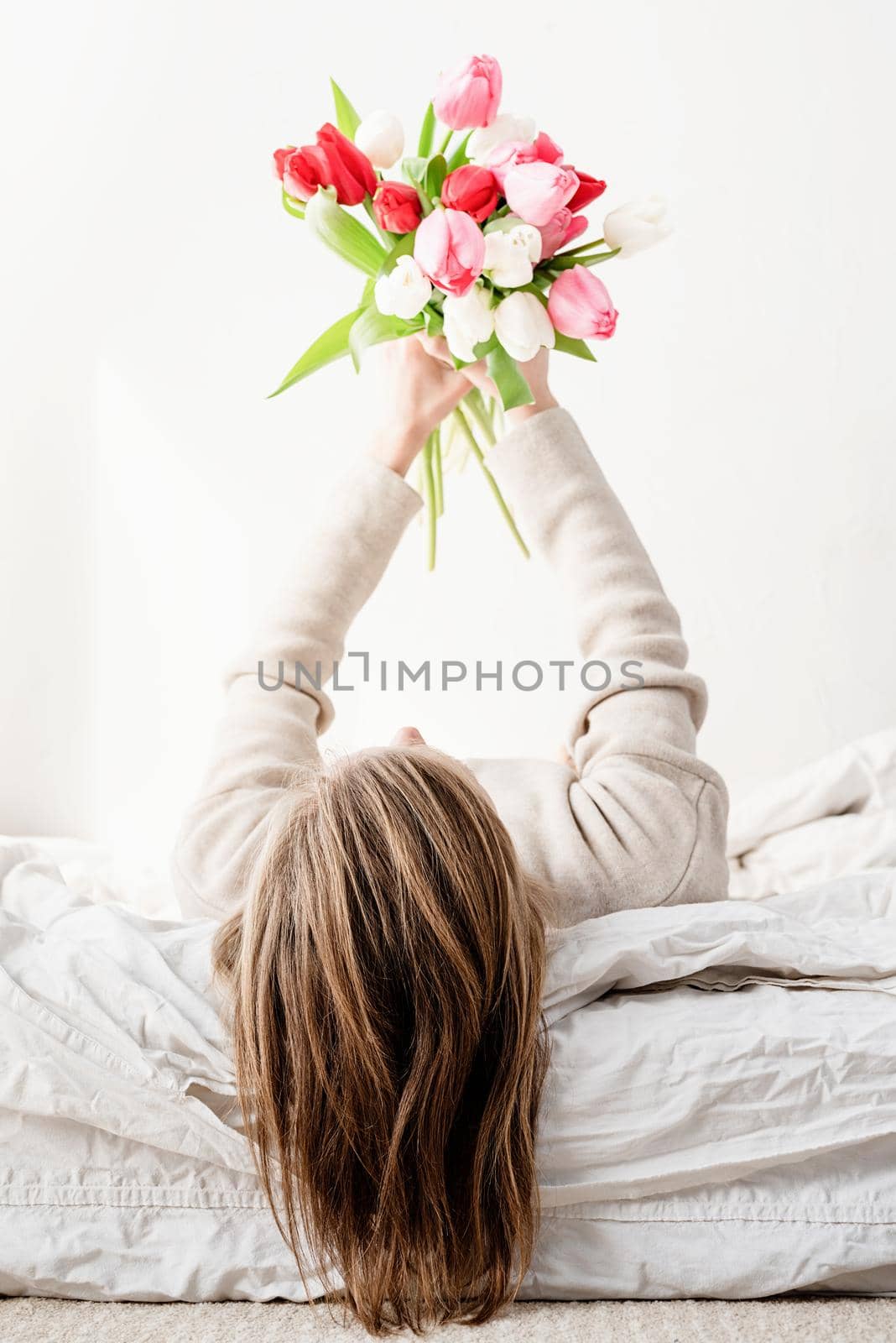 Happy woman lying on the bed wearing pajamas holding bright tulip flowers bouquet in outstretched hands