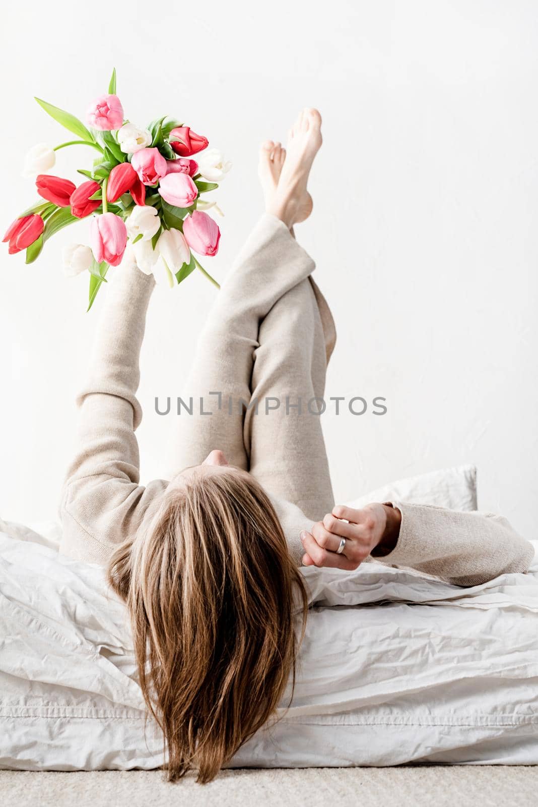 Happy woman lying on the bed wearing pajamas holding bright tulip flowers bouquet in outstretched hands
