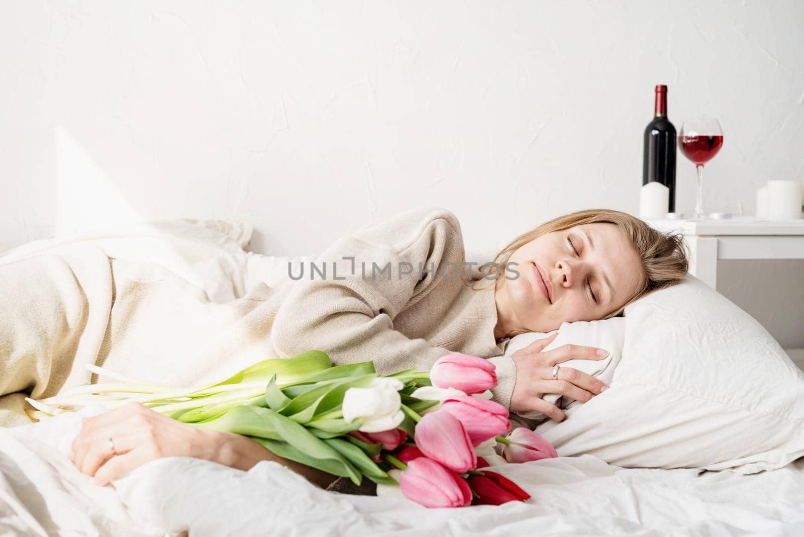 Happy young woman lying in the bed wearing pajamas holding tulip flowers bouquet