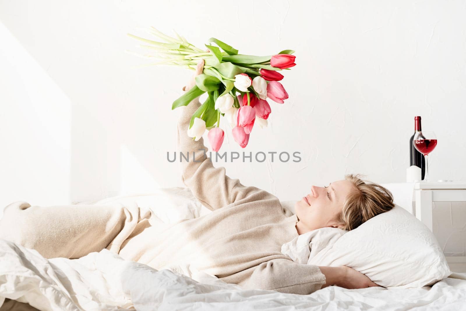 Happy young woman lying in the bed wearing pajamas holding tulip flowers bouquet