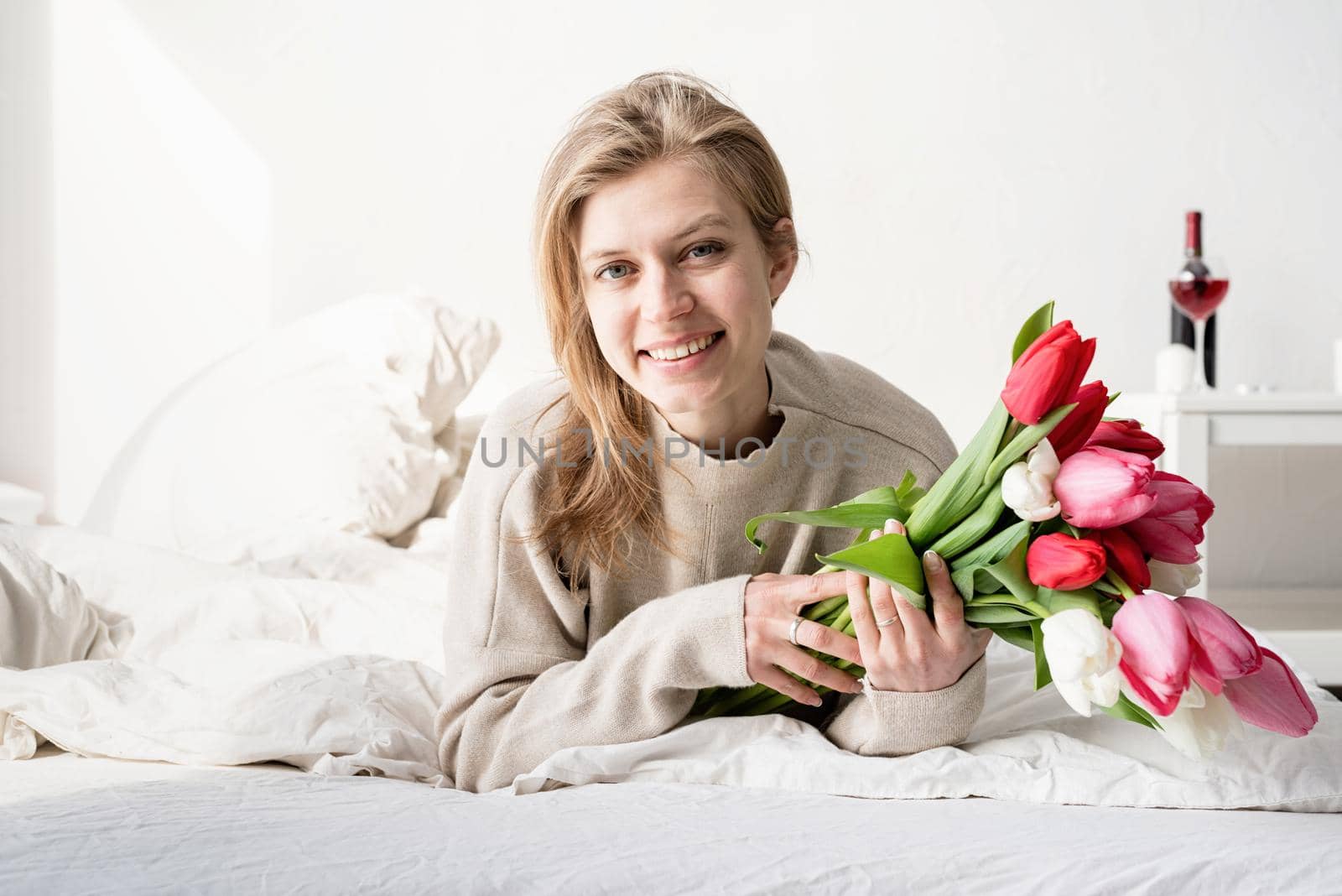 Happy young woman lying in the bed wearing pajamas holding tulip flowers bouquet