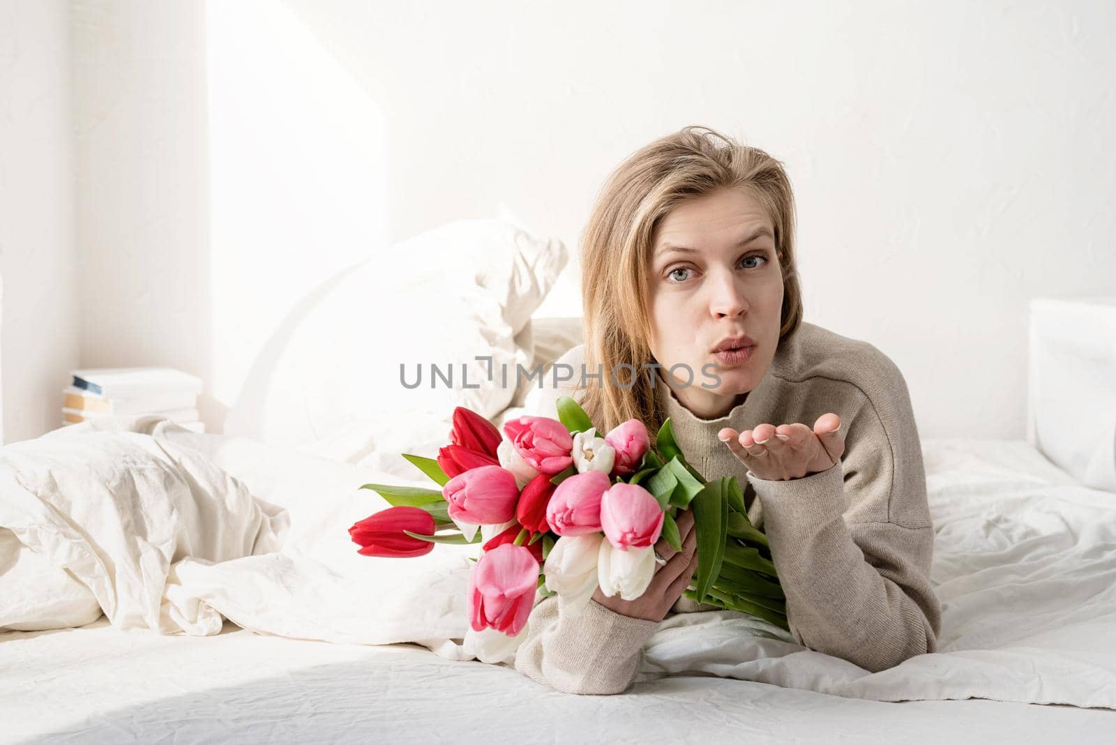 Happy young woman lying in the bed wearing pajamas holding tulip flowers bouquet and blowing a kiss