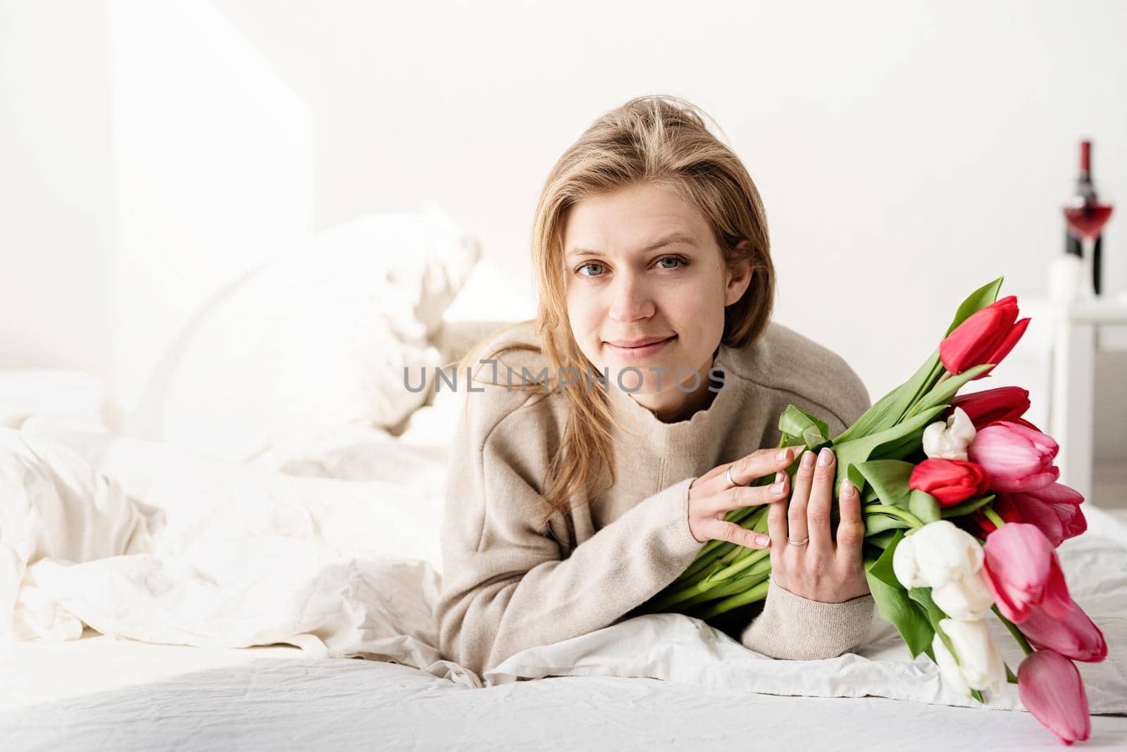 Happy woman lying in the bed wearing pajamas holding tulip flowers bouquet by Desperada