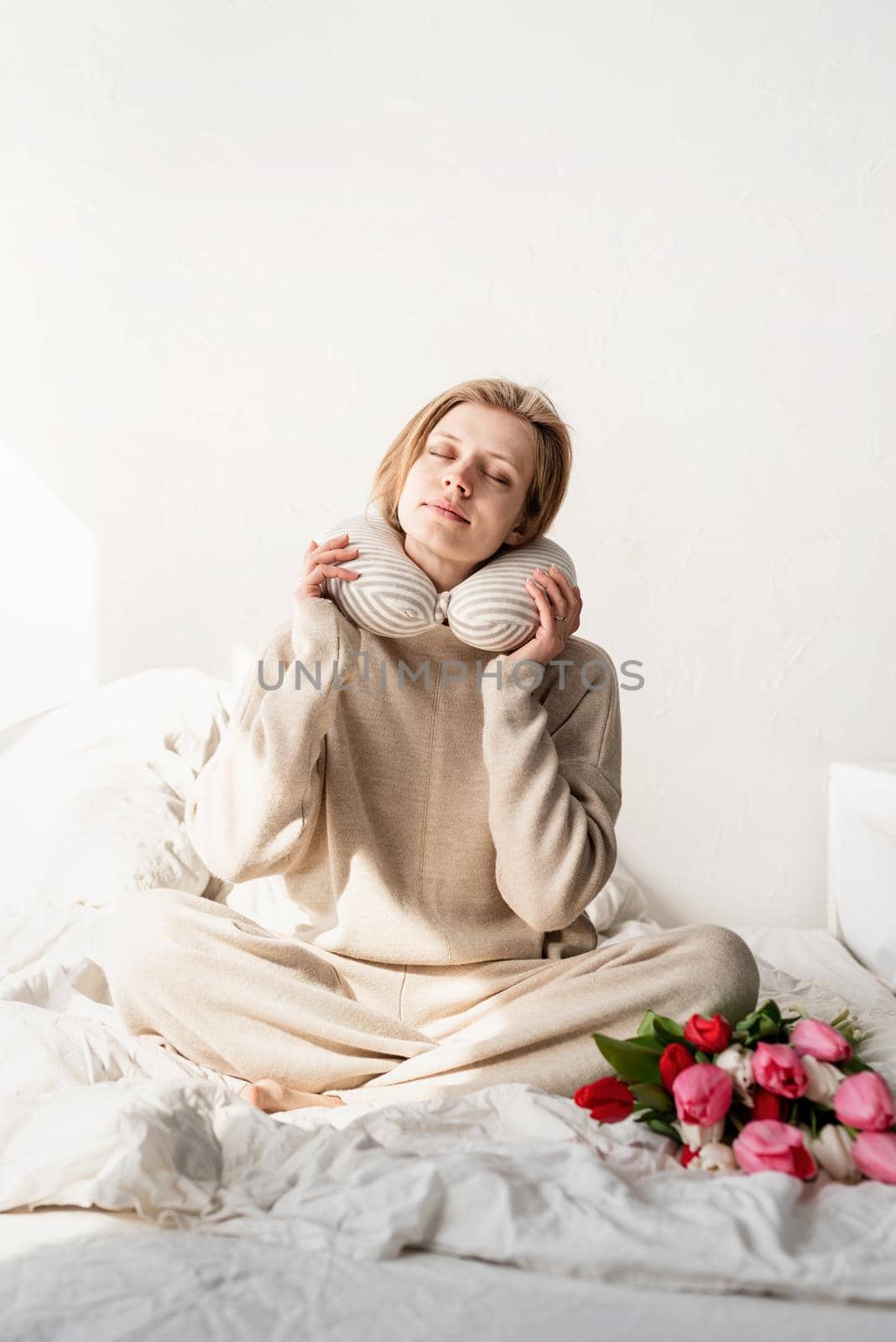 Sleepy woman sitting in bed wearing pajamas and pillow over the neck, tulip flowers on the bed