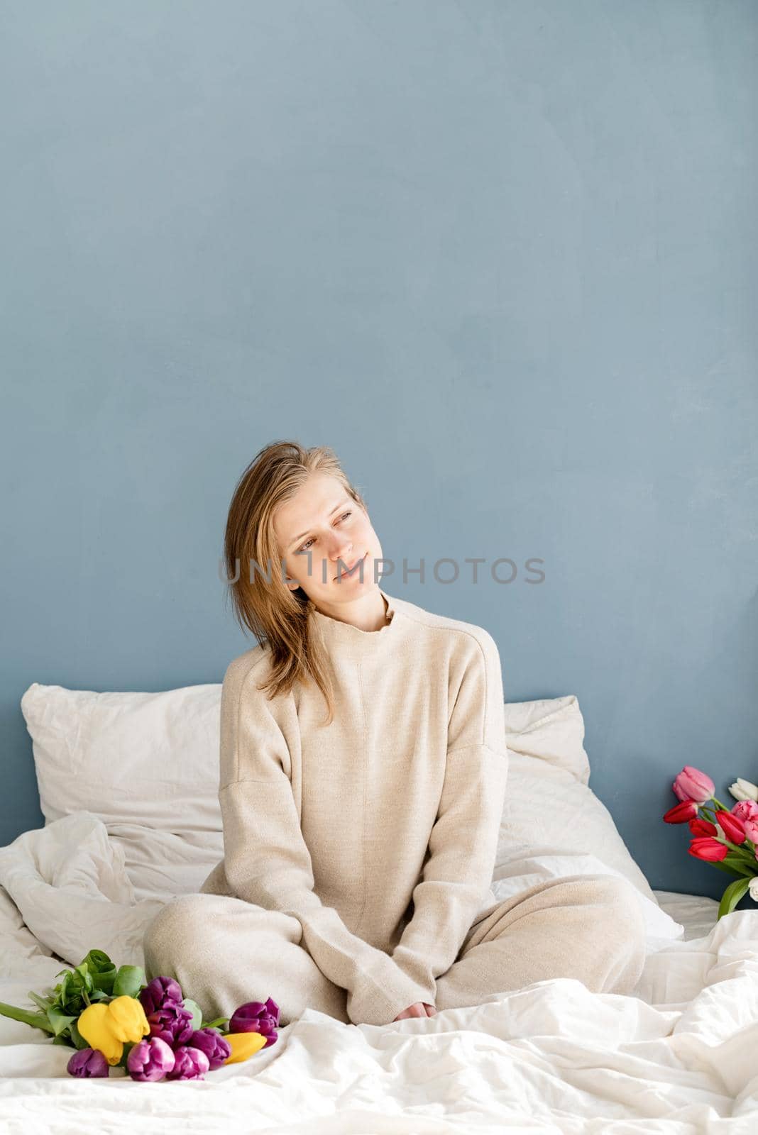 Happy woman sitting on the bed wearing pajamas holding tulip flowers bouquet, blue wall background by Desperada