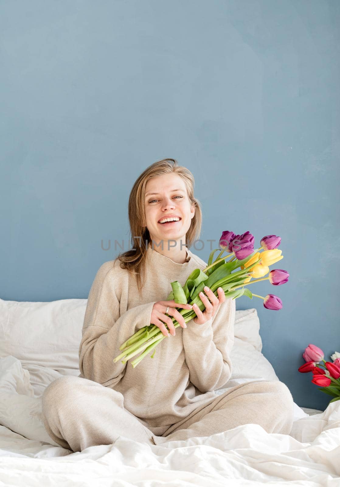 Happy woman sitting on the bed wearing pajamas holding tulip flowers bouquet, blue wall background by Desperada