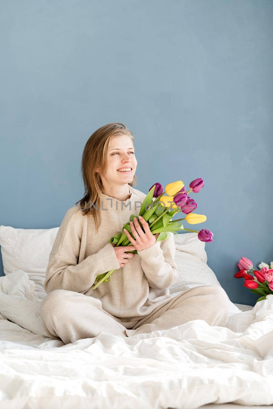 Happy woman sitting on the bed wearing pajamas holding tulip flowers bouquet, blue wall background by Desperada