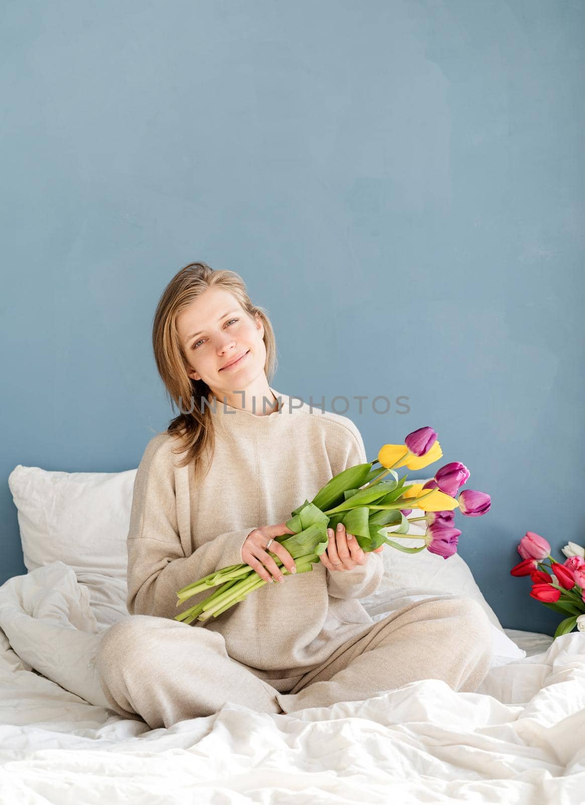 Happy woman sitting on the bed wearing pajamas holding tulip flowers bouquet, blue wall background by Desperada