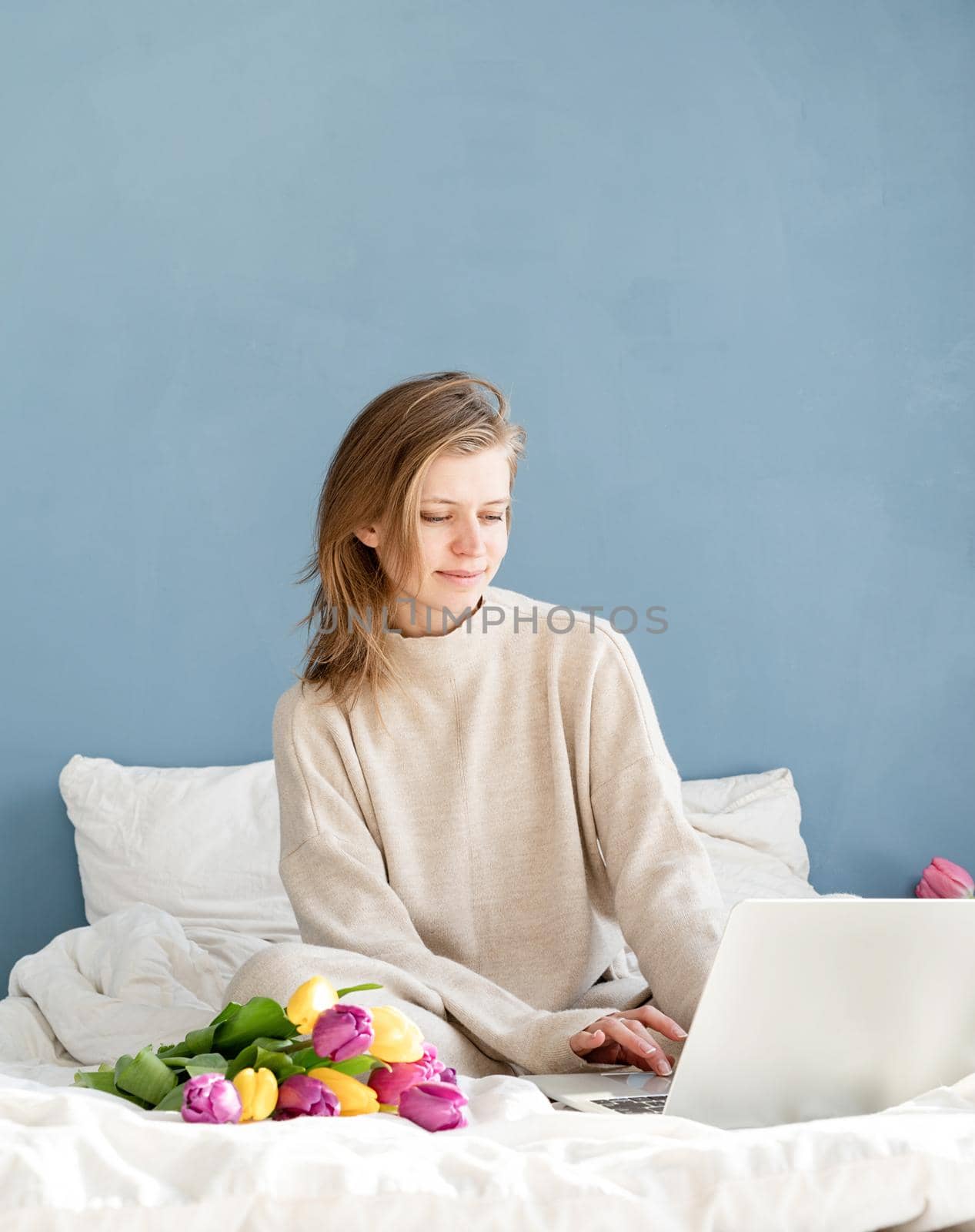 Happy smiling woman sitting on the bed wearing pajamas, with pleasure enjoying flowers, working on laptop
