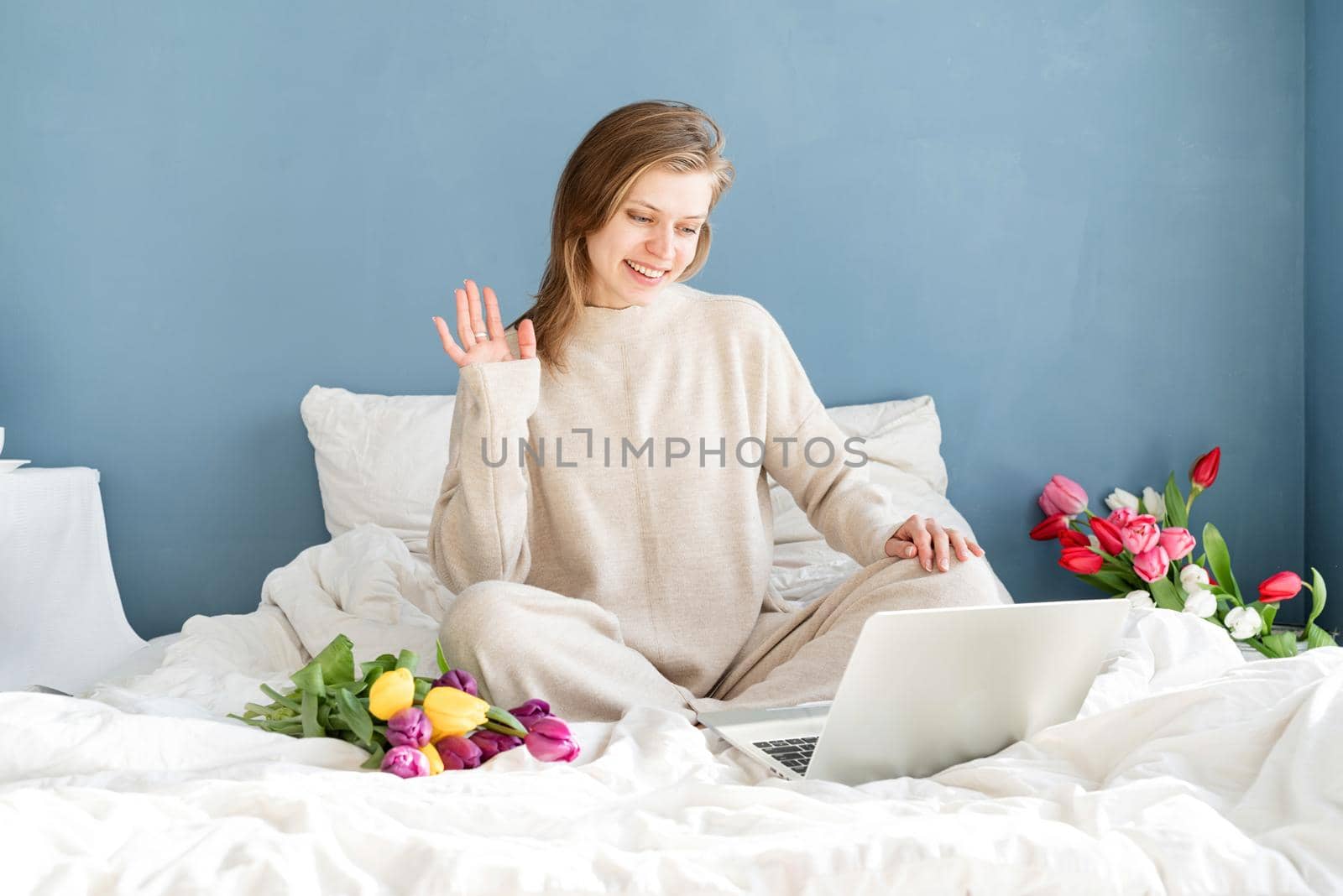 Happy smiling woman sitting on the bed wearing pajamas, with pleasure enjoying flowers, chatting using laptop