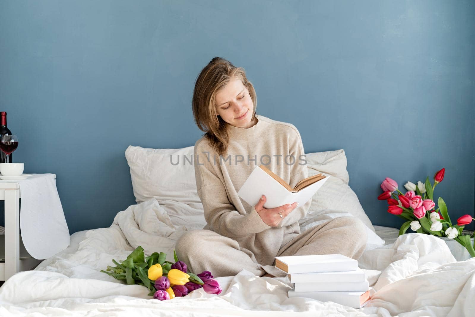 Happy smiling woman sitting on the bed wearing pajamas, with pleasure enjoying flowers and reading a book