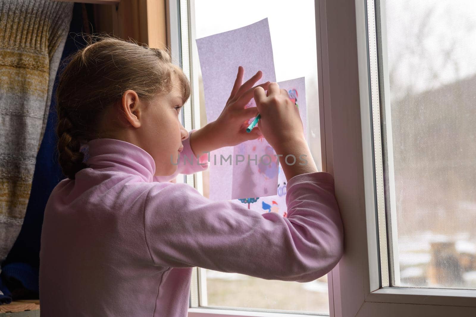 The girl copies the printed picture onto a blank sheet by attaching the picture to the window glass