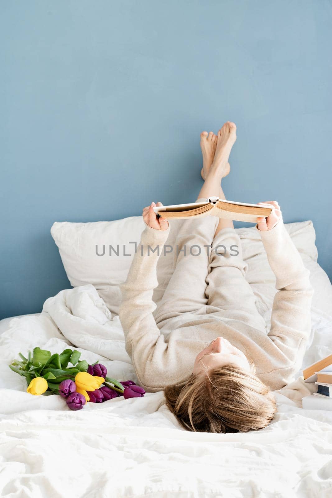 Happy smiling woman sitting on the bed wearing pajamas, with pleasure enjoying flowers and reading a book