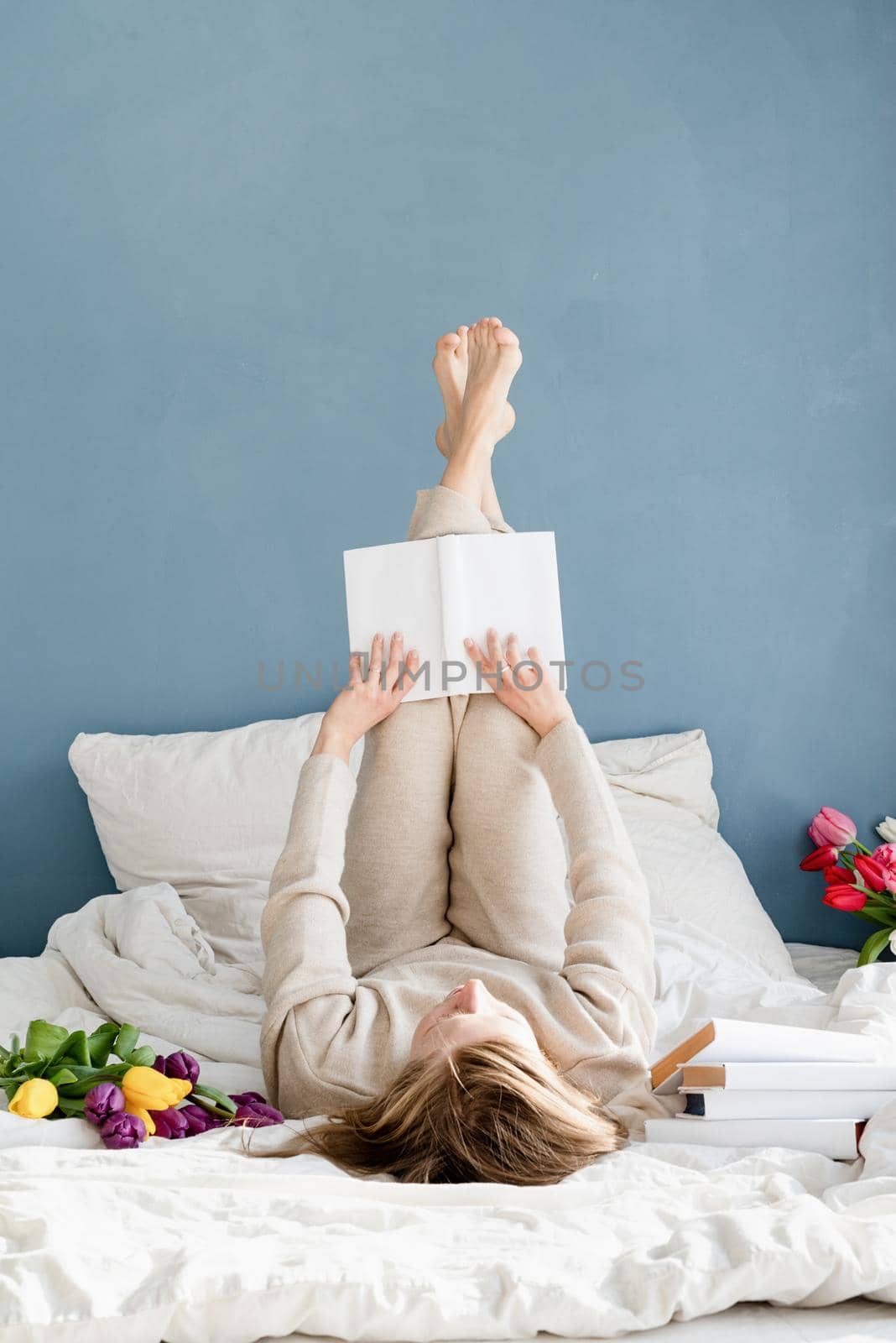 Happy smiling woman lying on the bed wearing pajamas, with pleasure enjoying flowers and reading a book