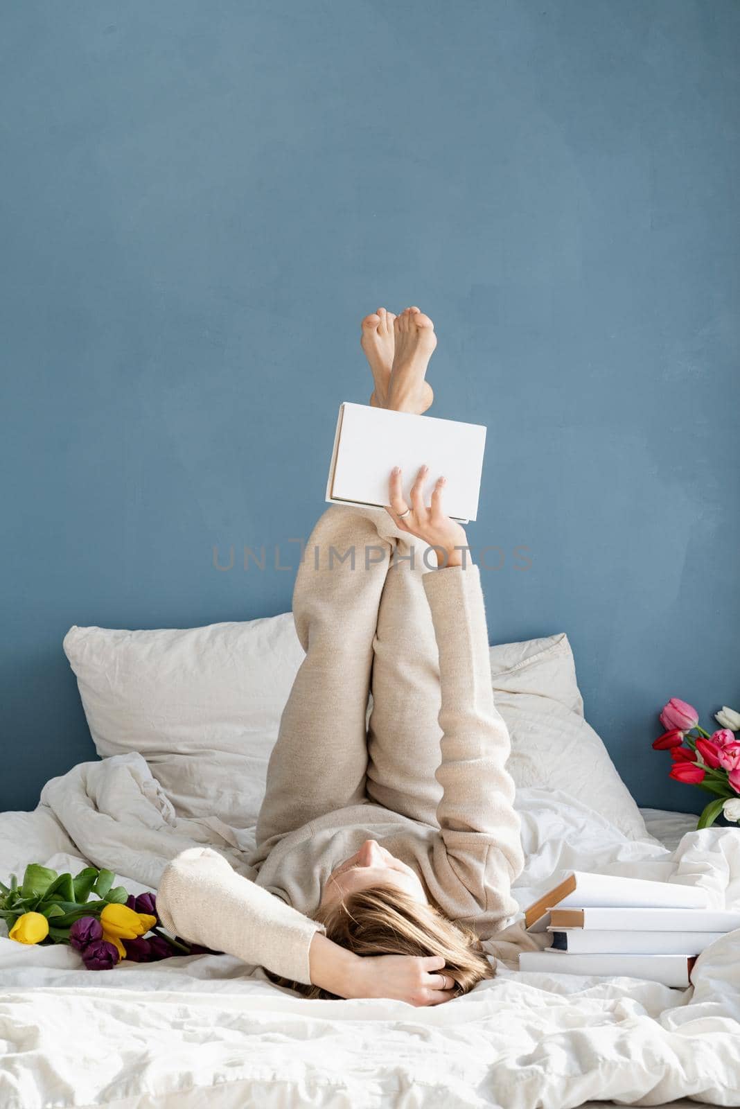 Happy smiling woman lying on the bed wearing pajamas, with pleasure enjoying flowers and reading a book