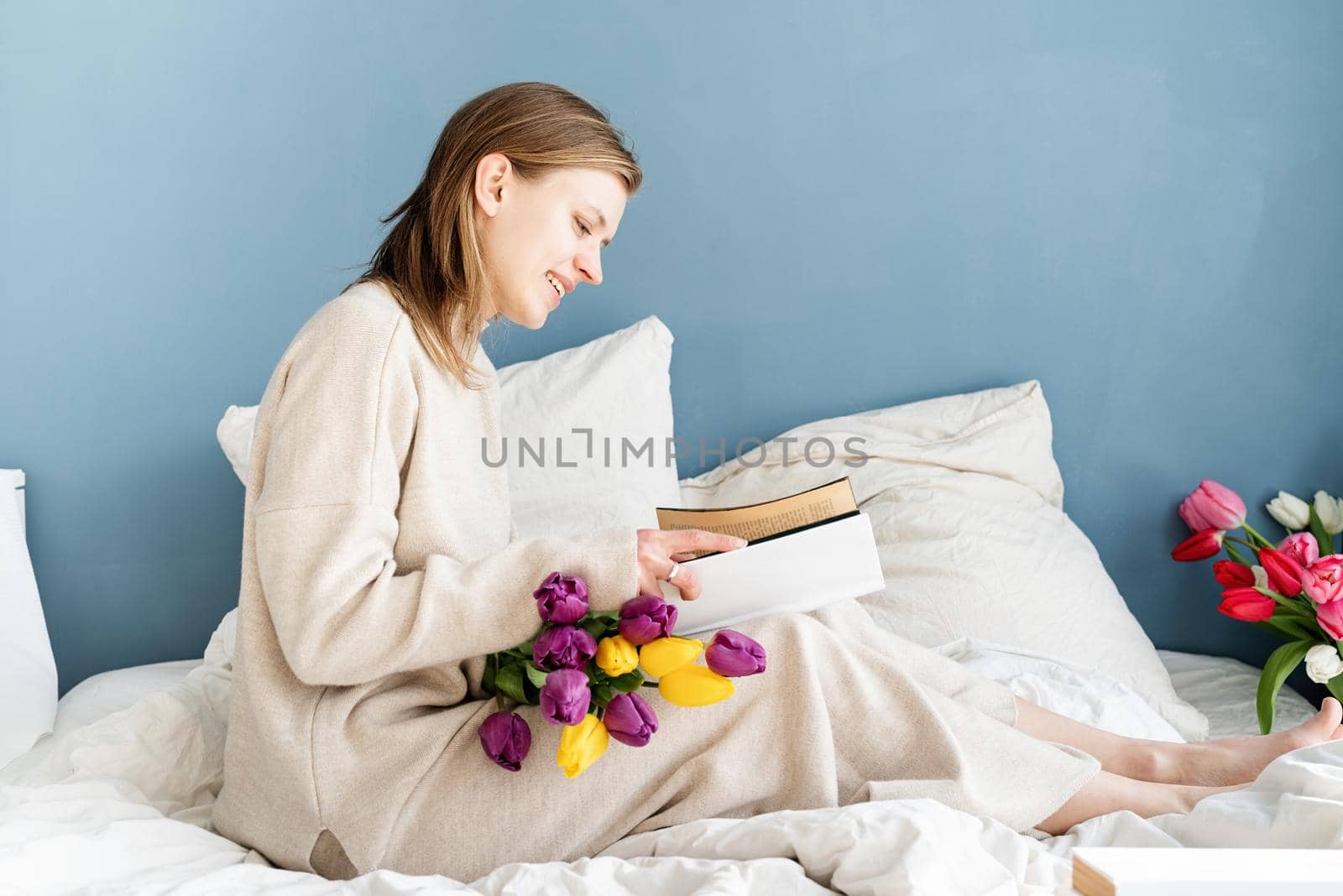 Happy woman sitting on the bed wearing pajamas reading a book by Desperada