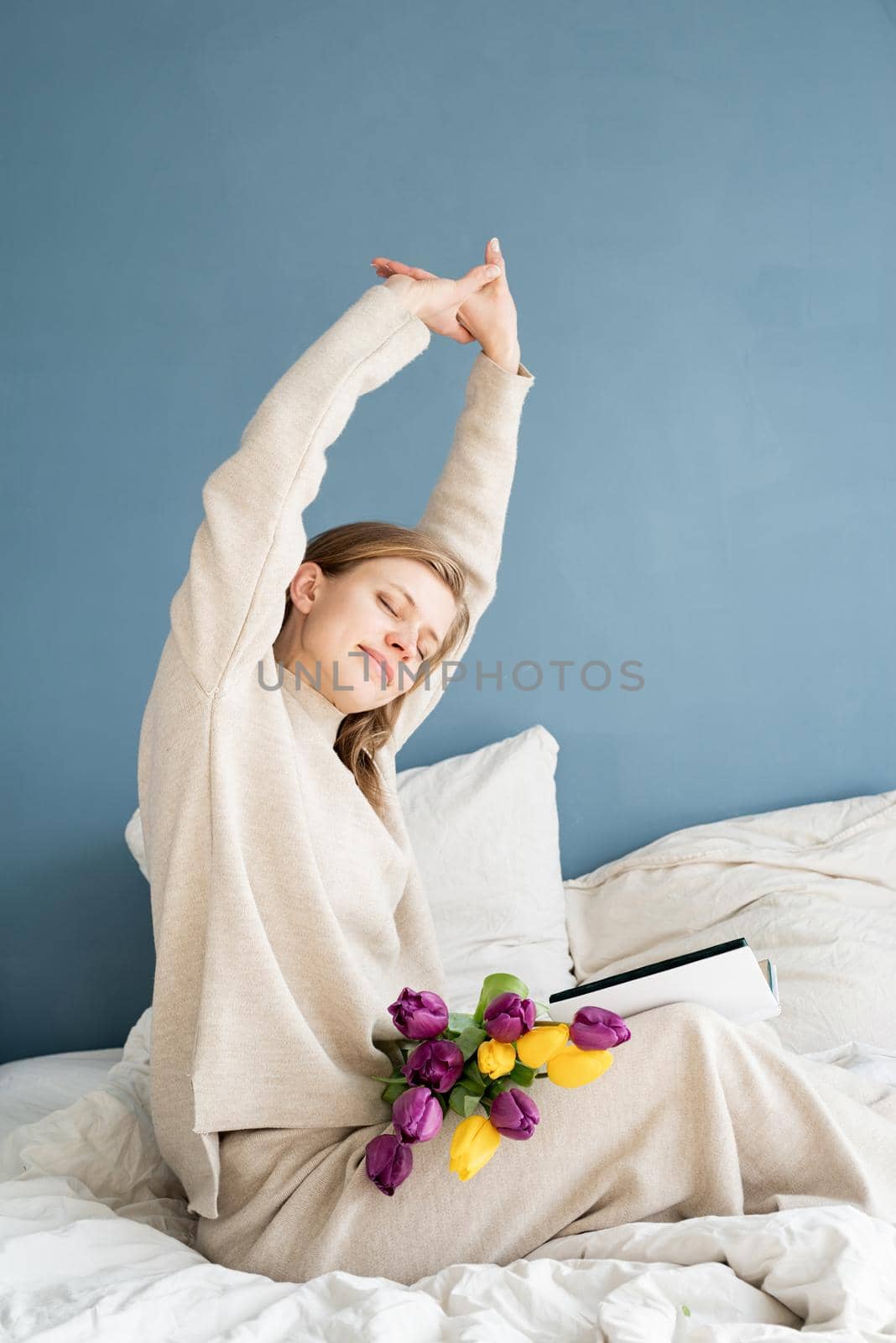 Happy woman sitting on the bed wearing pajamas stretching reading a book by Desperada