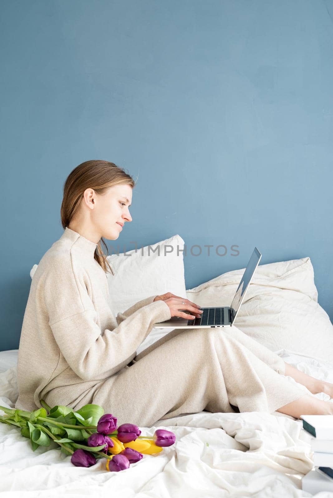 Happy woman sitting on the bed wearing pajamas holding tulip flowers bouquet chatting on laptop by Desperada