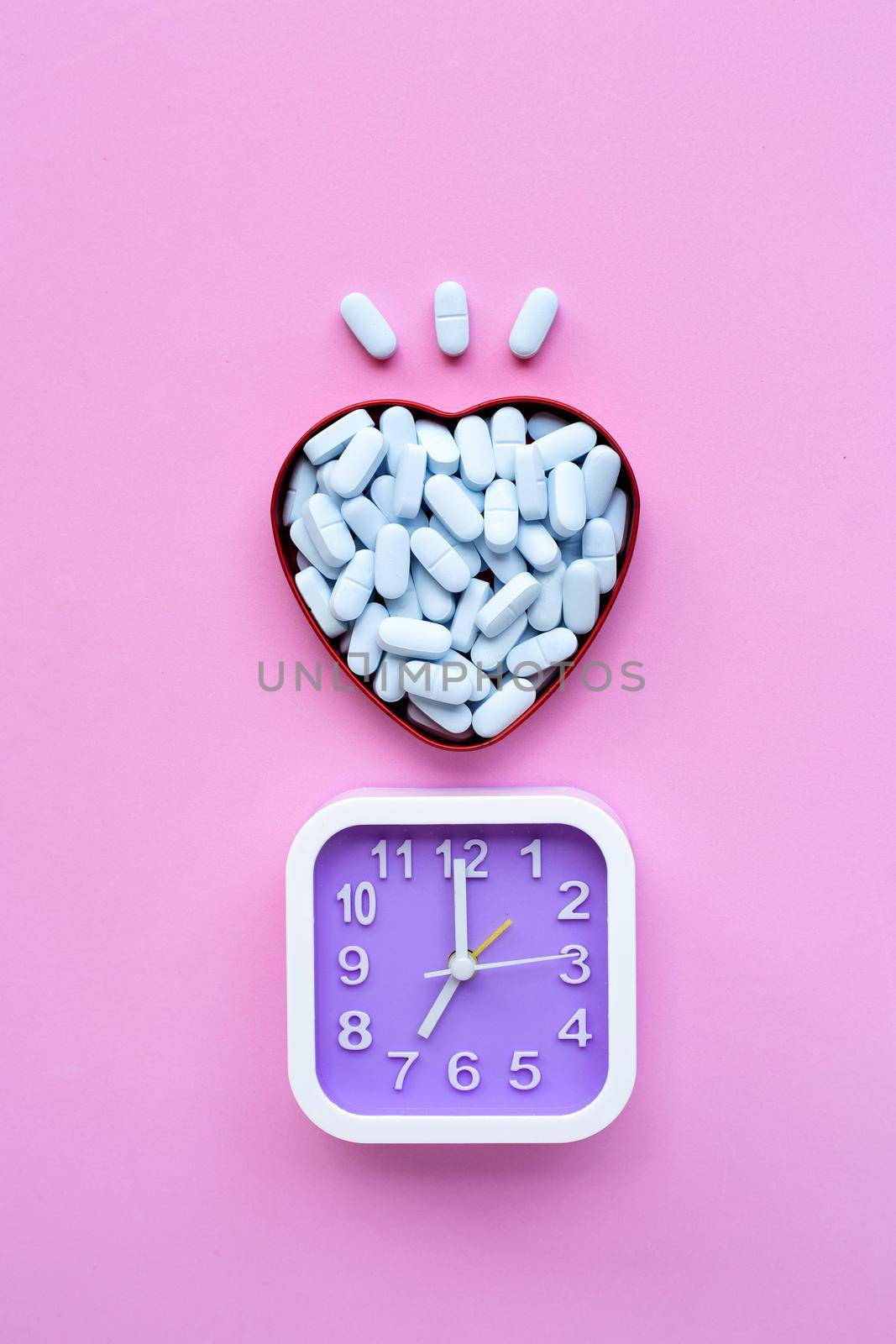 Clock with medical blue pills in heart shaped box on pink background. Top view