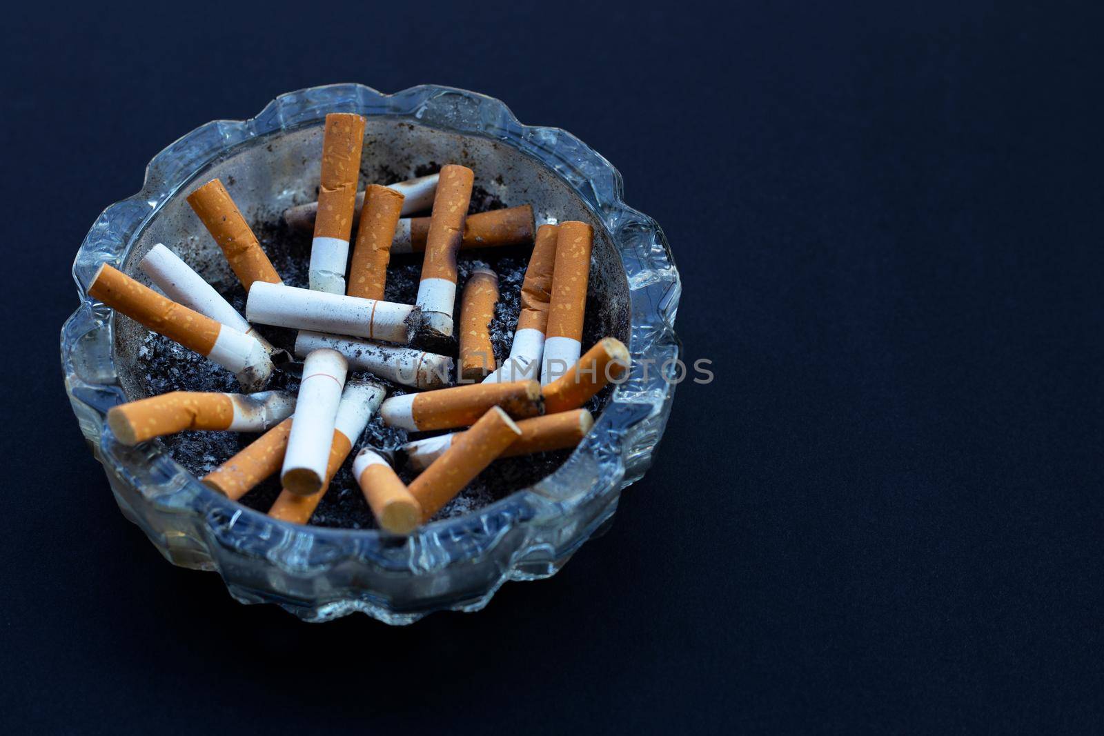 Cigarettes buds in a transparent ashtray on dark background.